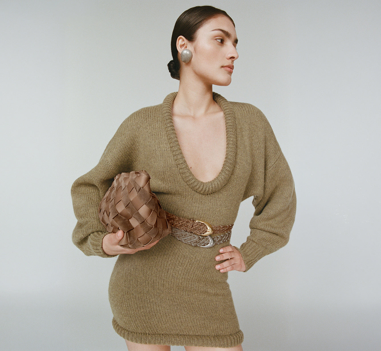A woman models an olive green sweater dress with a deep V-neck, paired with a brown handwoven suede and neoprene belt and holding a brown handwoven suede and neoprene clutch against a light gray studio background.