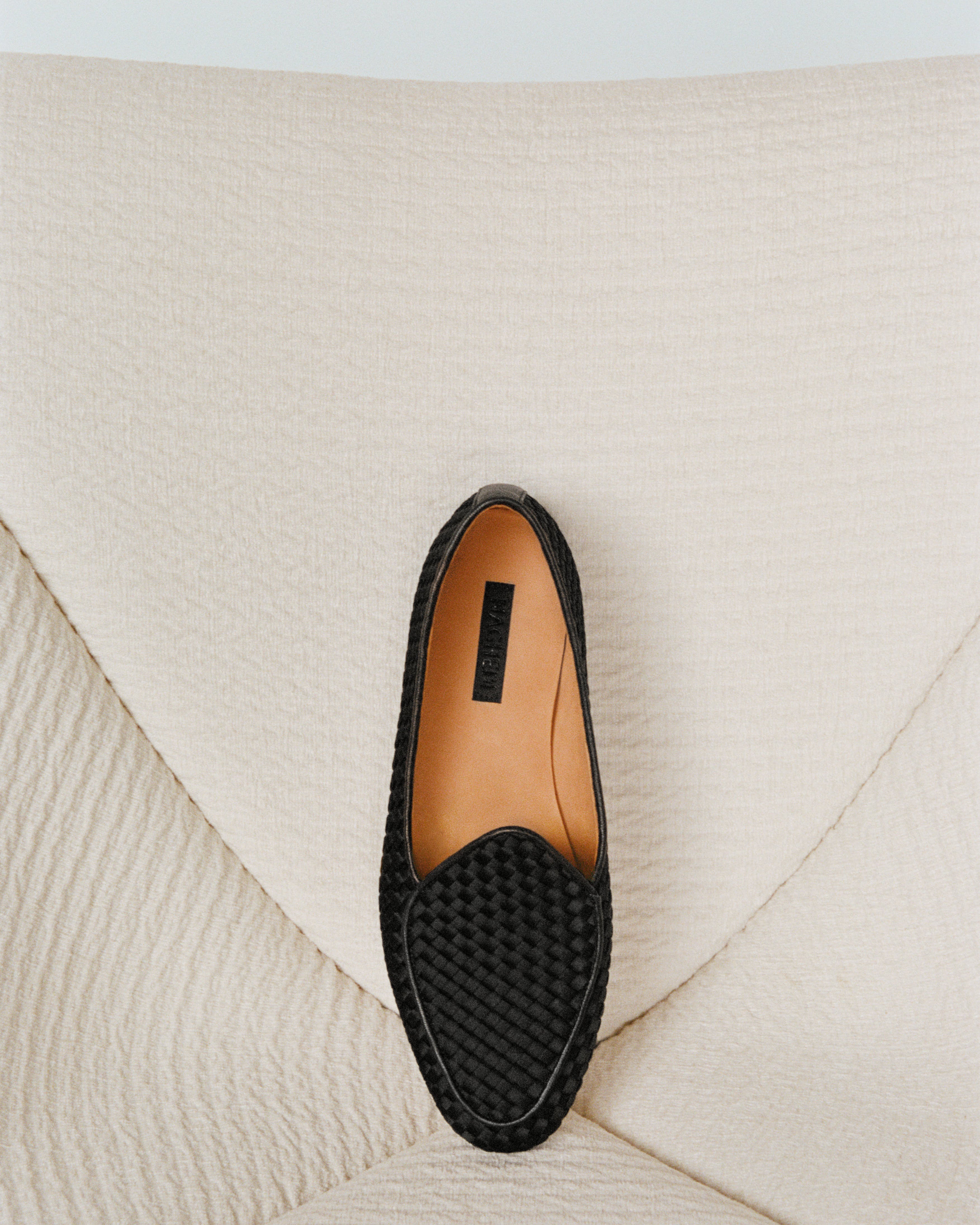 A single black handwoven satin loafer with leather trim shown from above on a cream colored couch
