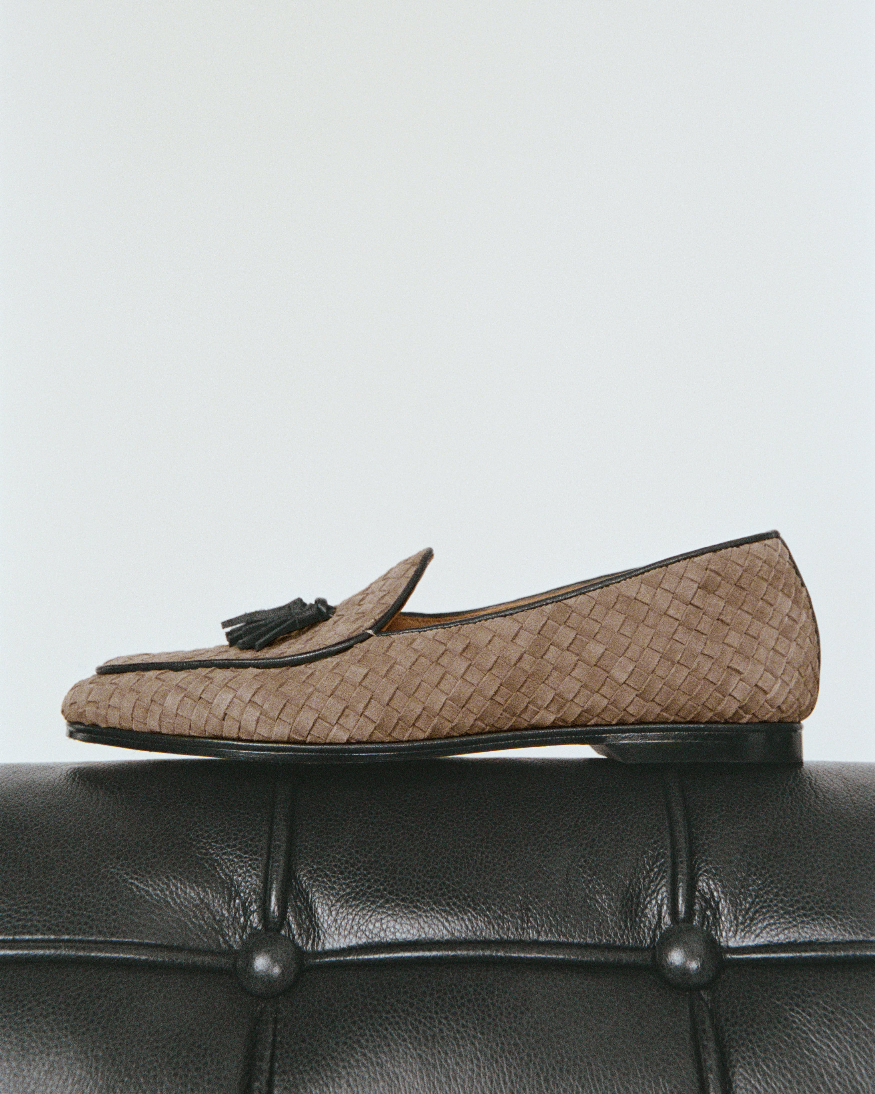 A side view of a suede loafer in beige with a black tassel and black trim on top of a black leather couch.