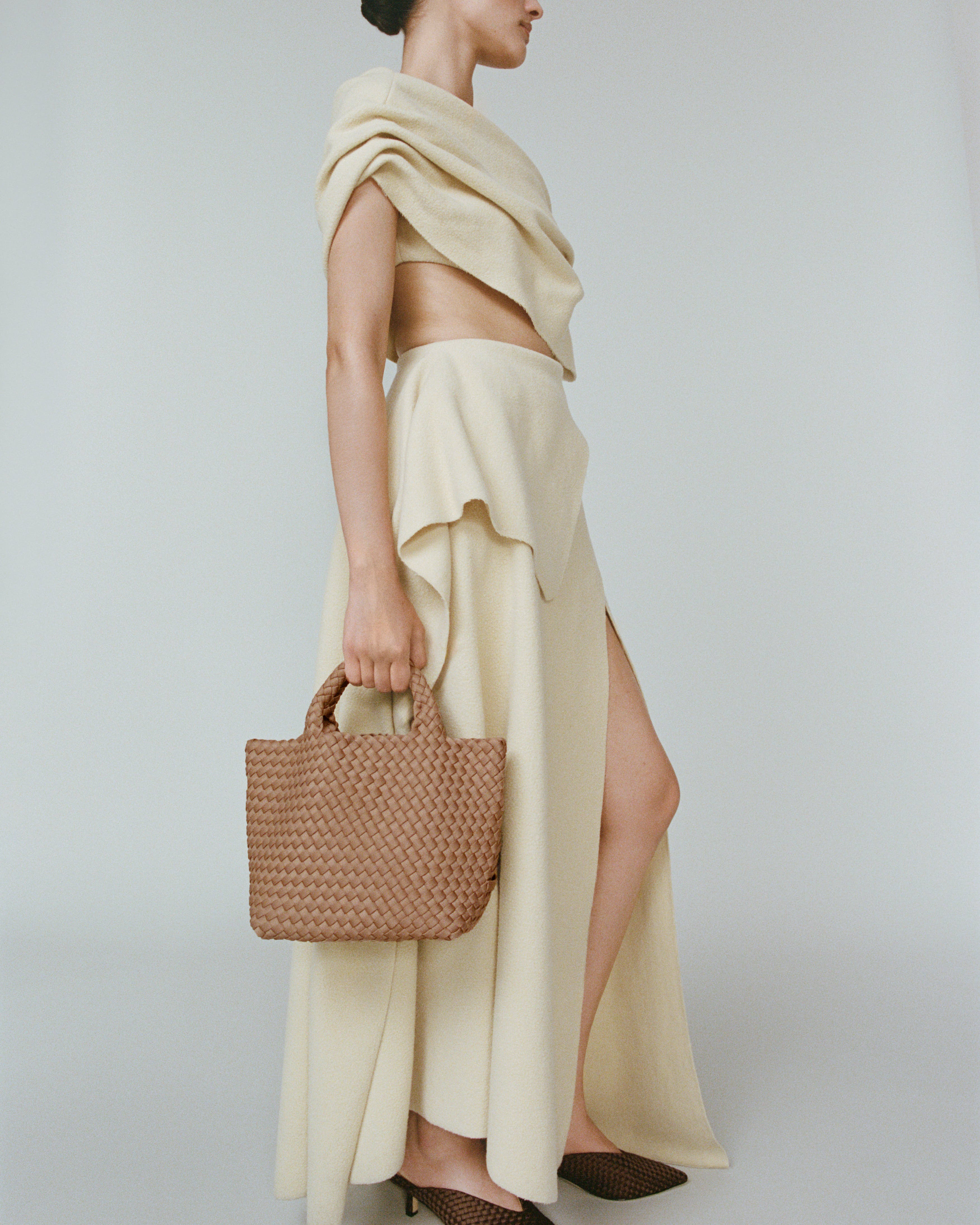 A model in a side profile wearing a cream-colored dress, holding a St. Barths small tote bag in Seychelles at her side