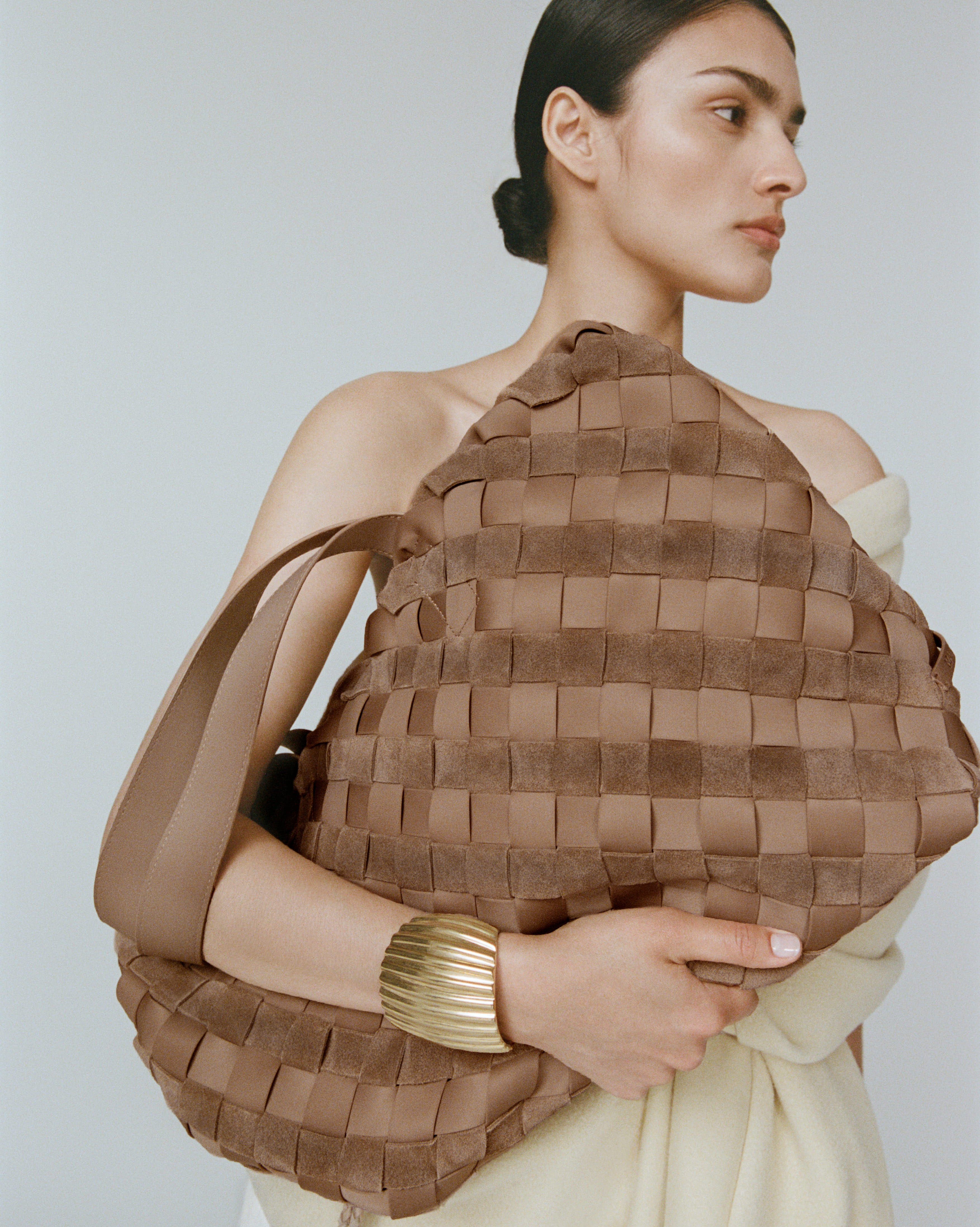 A woman in a cream off-the-shoulder dress holds a Medium Como handwoven light brown Tote in suede and neoprene fabric.