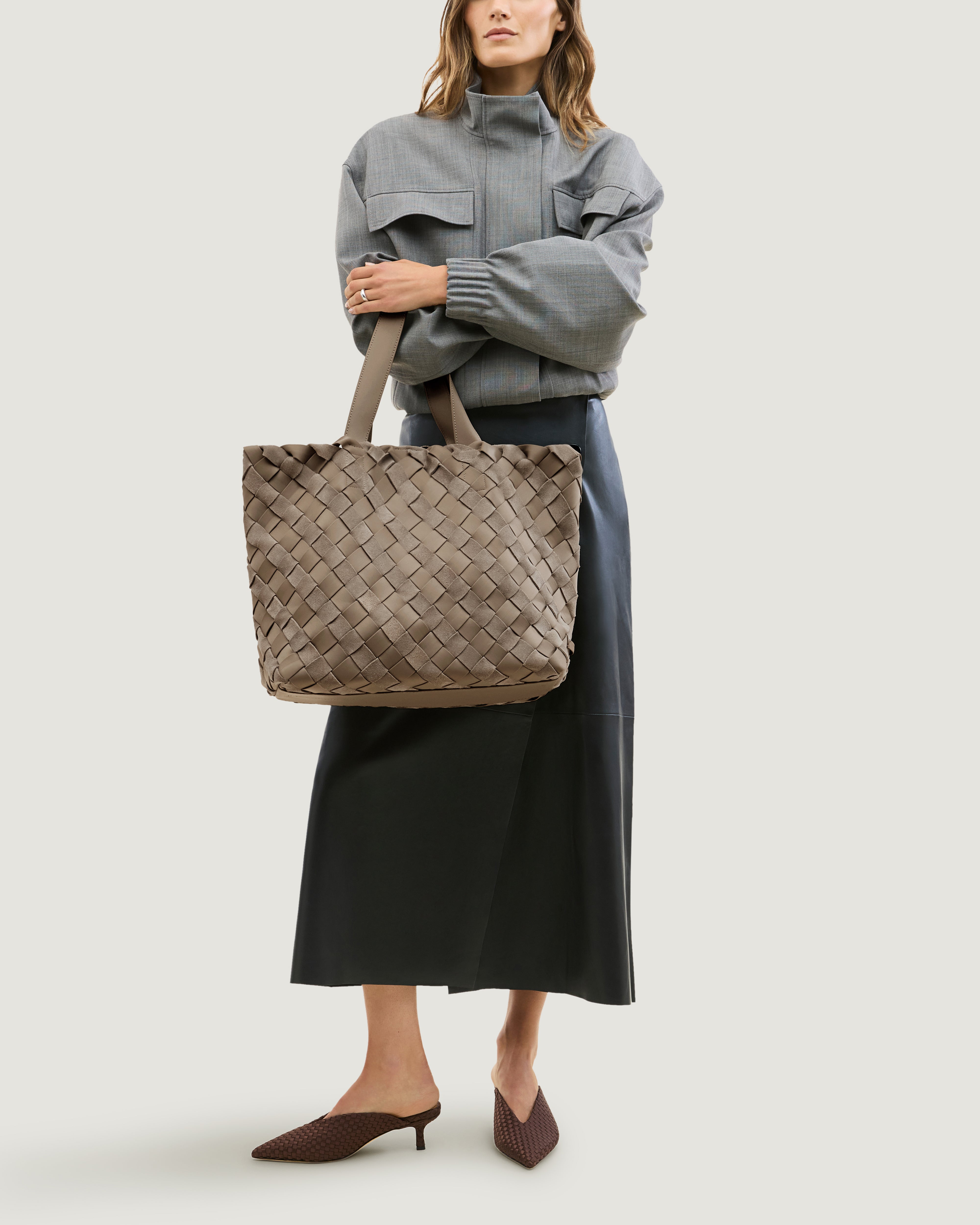 Model holding a large handwoven suede and neoprene tote bag in taupe brown with dual top handles in front of her body.