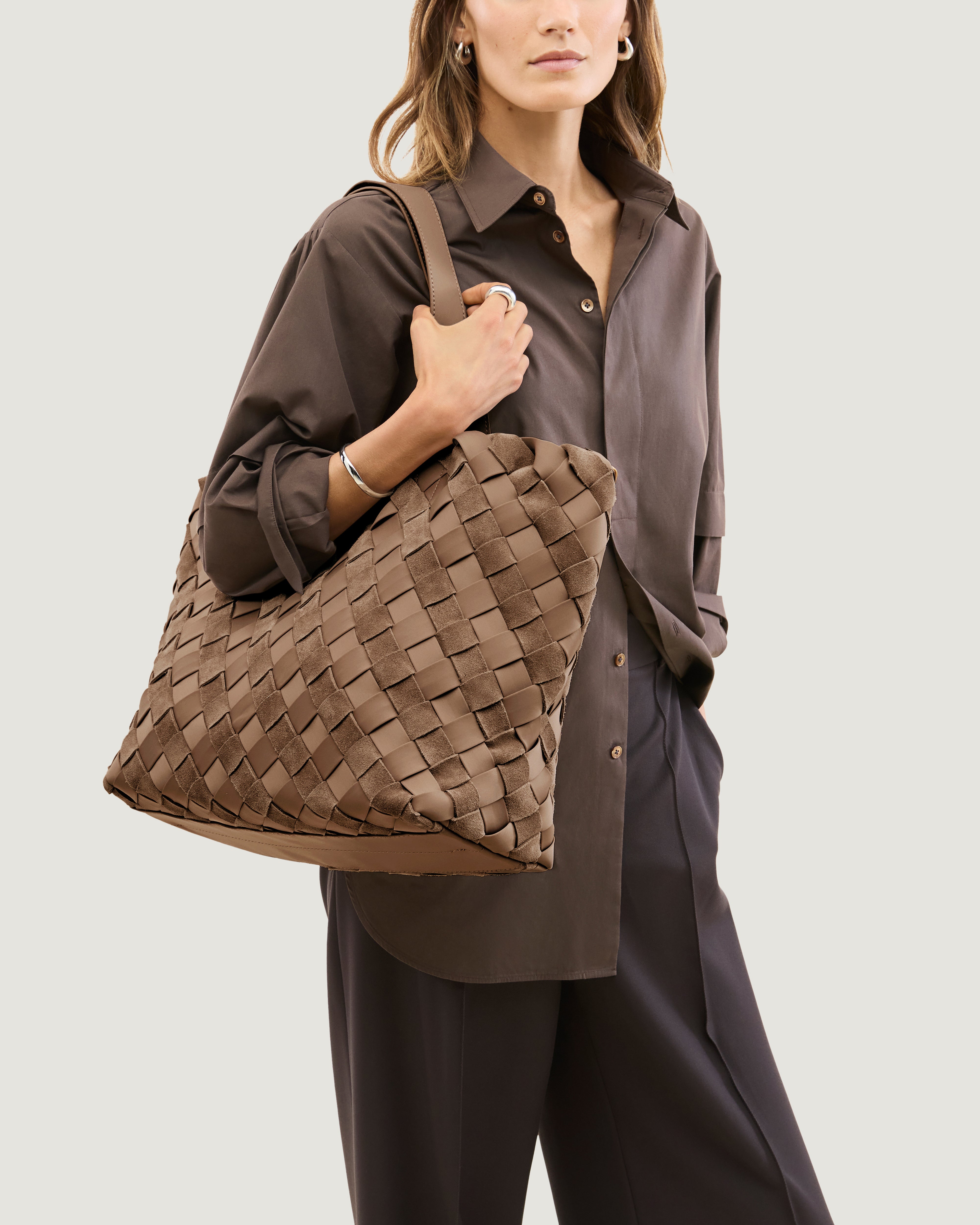A model carrying a handwoven suede and neoprene tote bag in a warm taupe-brown color, featuring a diamond-patterned weave and two flat shoulder straps over her shoulder.