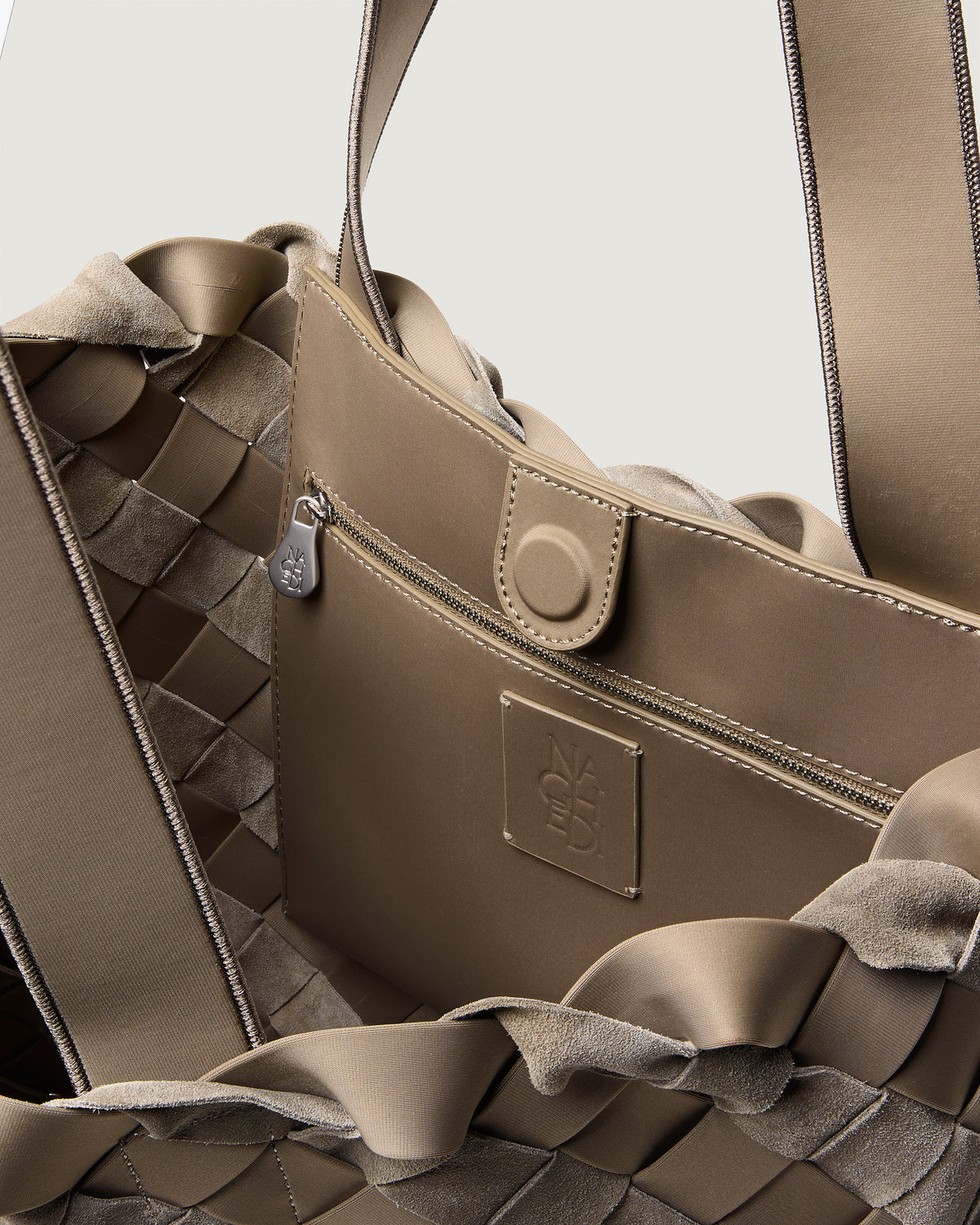 Close-up of a handwoven suede and neoprene shoulder bag in taupe-brown, showing the braided texture, a zip pocket, a snap tab, and a small embossed logo patch.