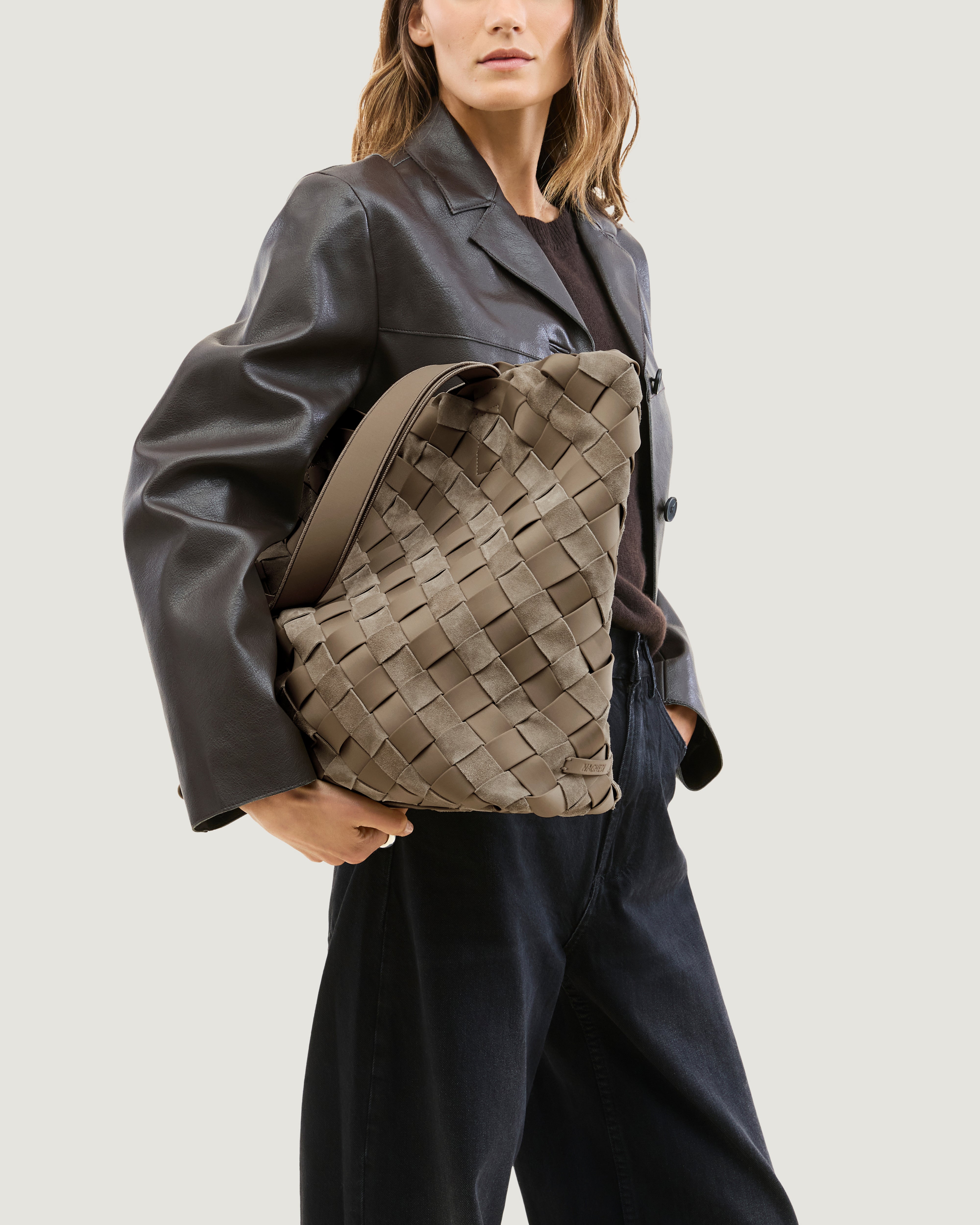 Model holding a handwoven suede and neoprene tote bag in a taupe-beige color, featuring a basketweave pattern and two matching shoulder straps under her arm.