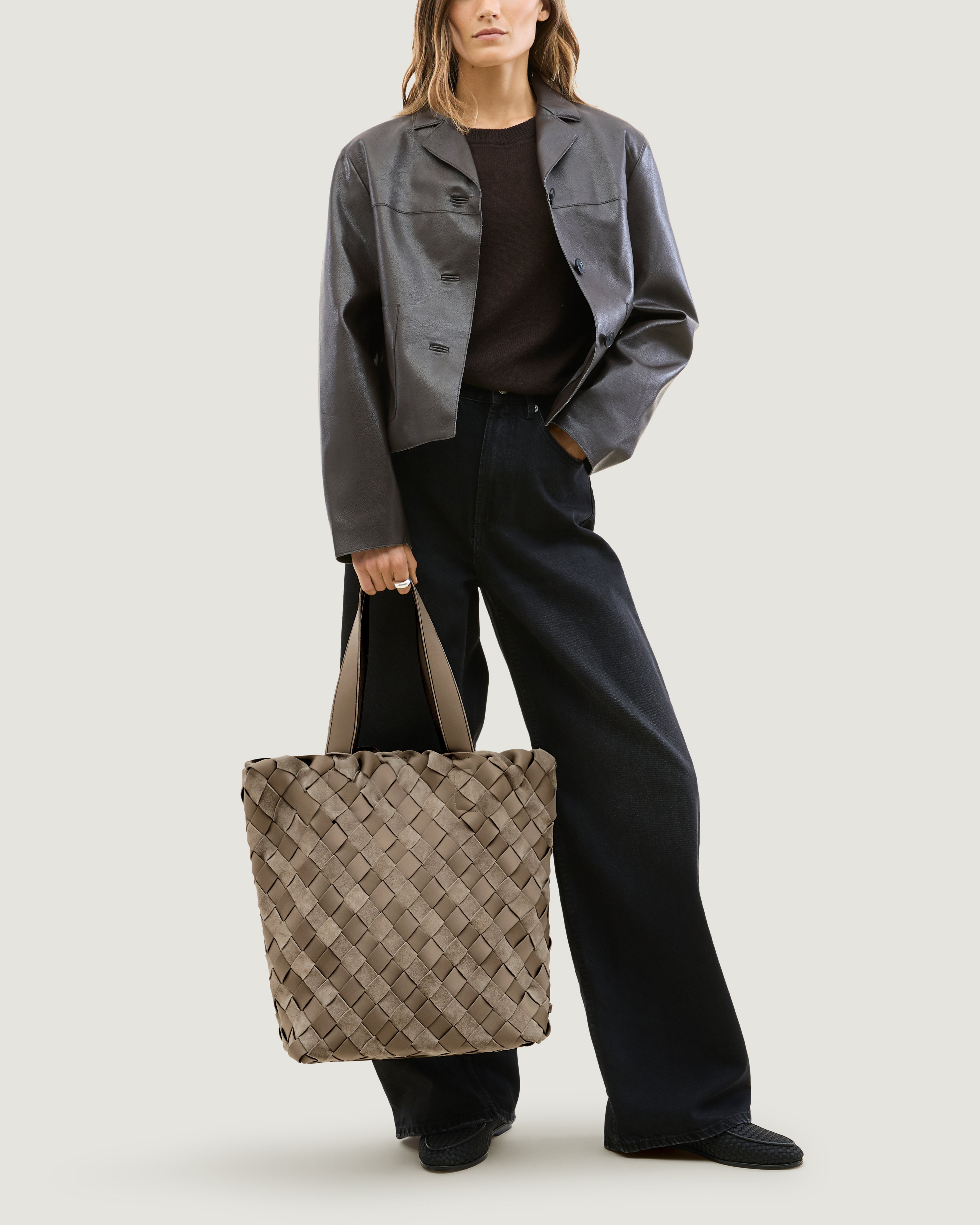 Model holding a handwoven suede and neoprene tote bag in a taupe-beige color, featuring a basketweave pattern and two matching shoulder straps at her side