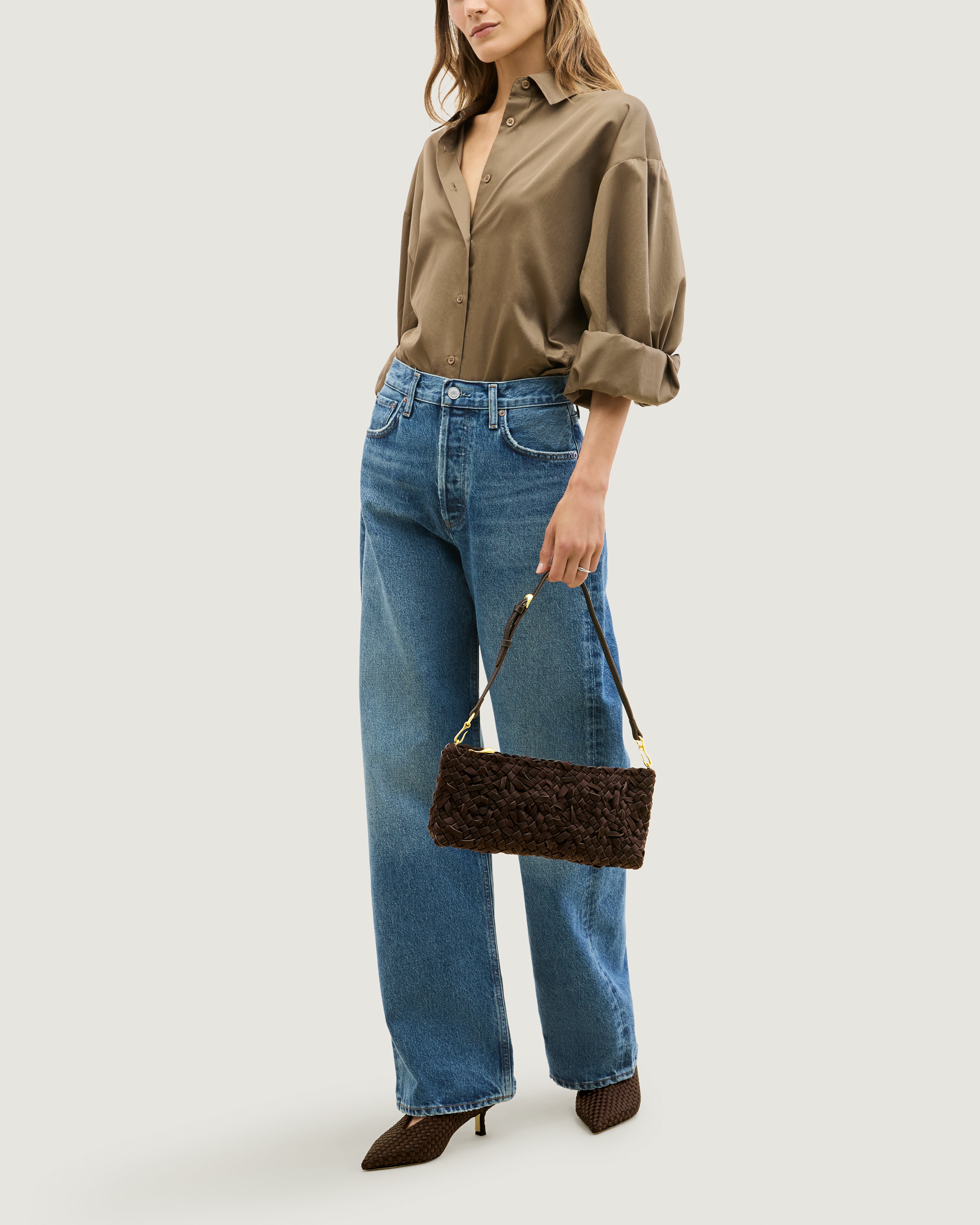 Model holding handwoven neoprene shoulder bag in chocolate brown color with a crosshatch nest weave pattern and gold hardware in front of her body.