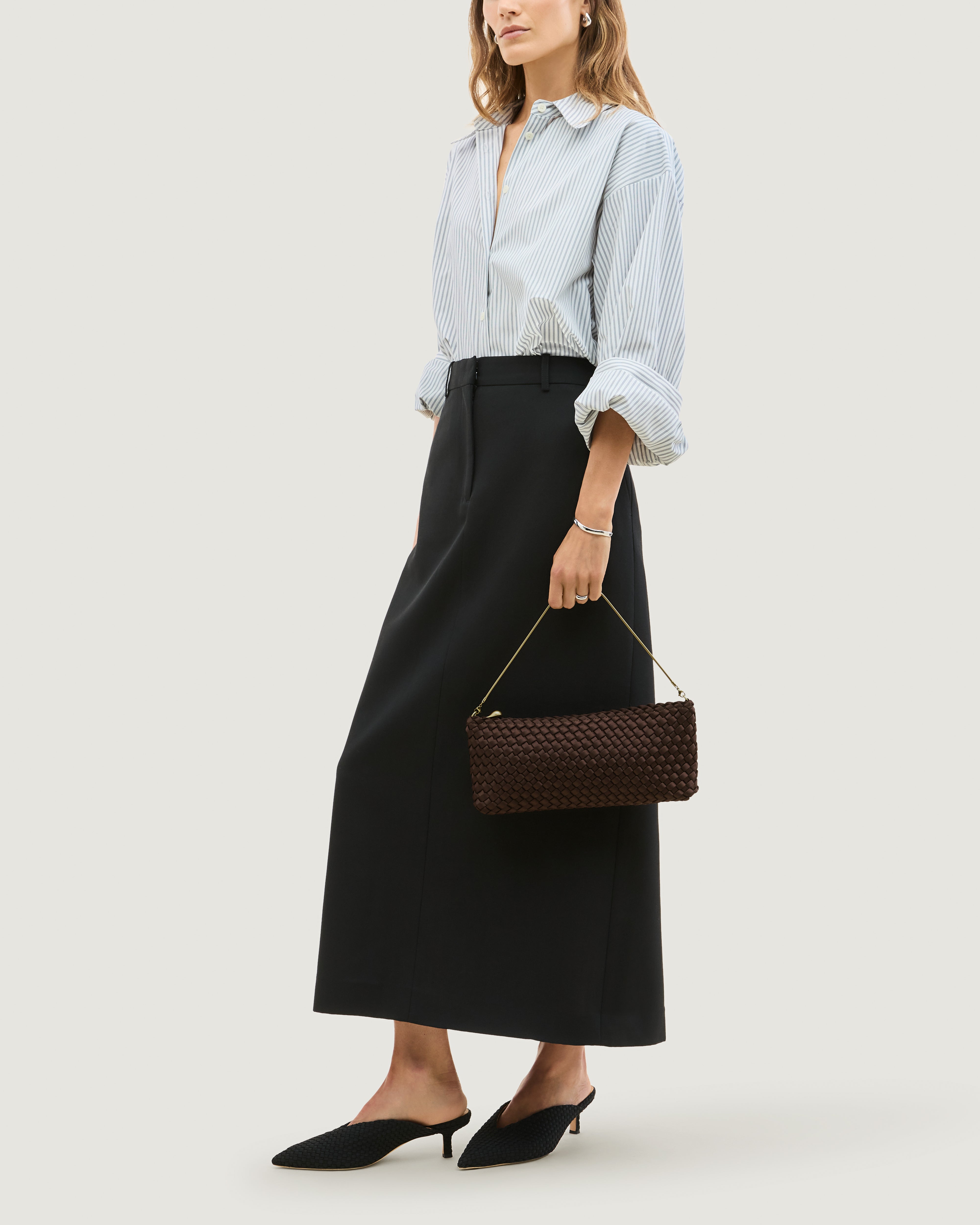 Model holding a handwoven satin neoprene shoulder bag in chocolate brown with a thin gold metal chain strap as a clutch in front of her body.