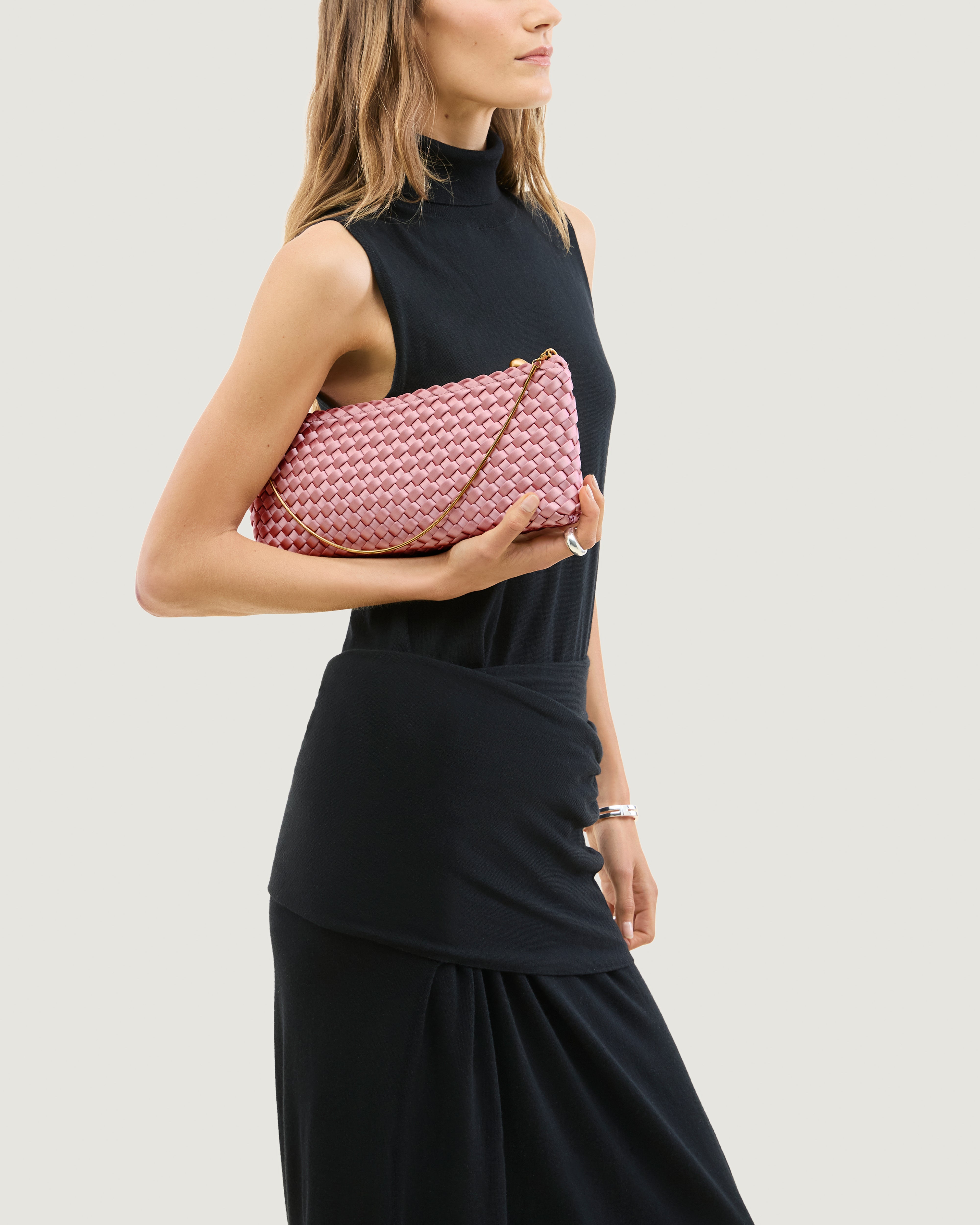 Model holding a handwoven satin neoprene shoulder bag in pink with a thin gold metal chain strap.
