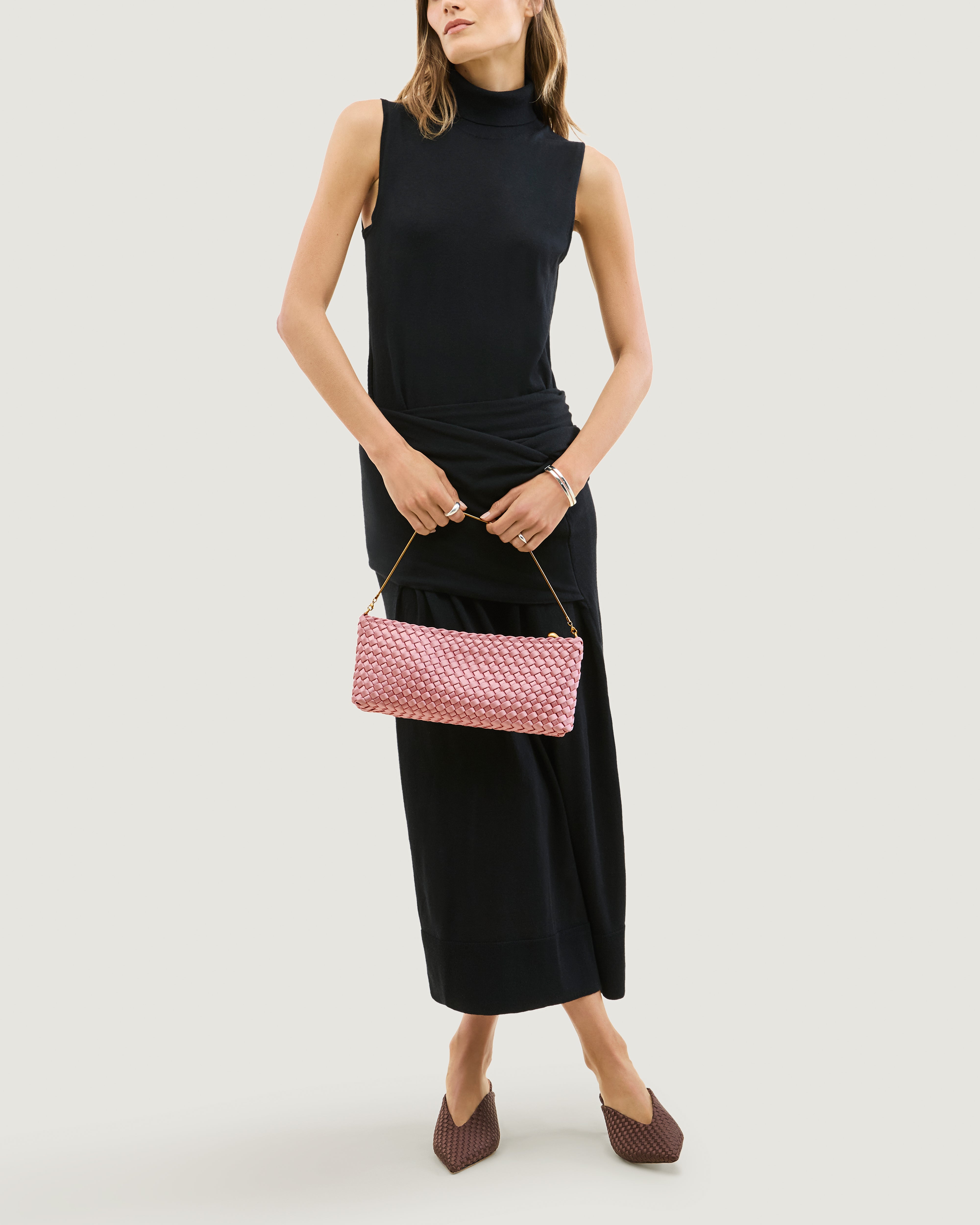 Model holding a handwoven satin neoprene shoulder bag in pink with a thin gold metal chain strap as a clutch in front of her body.