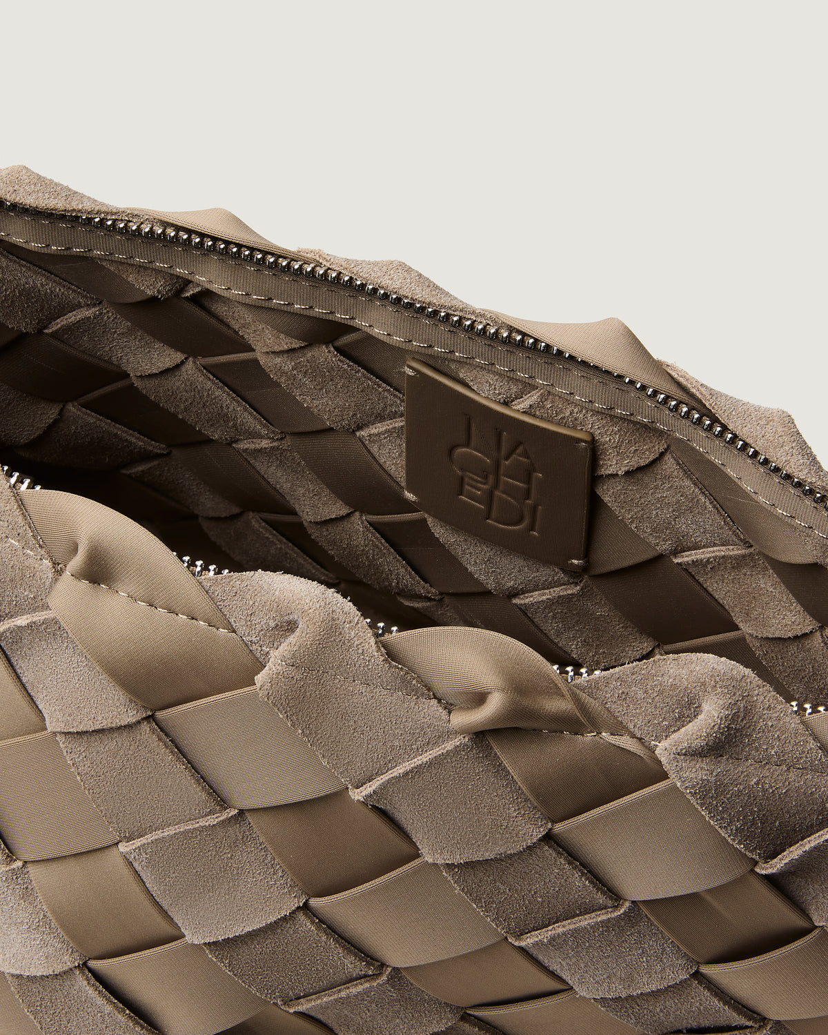 Close-up of a handwoven suede and neoprene bag interior in taupe-brown, showing a zippered opening, textured weave, and a small embossed logo patch.
