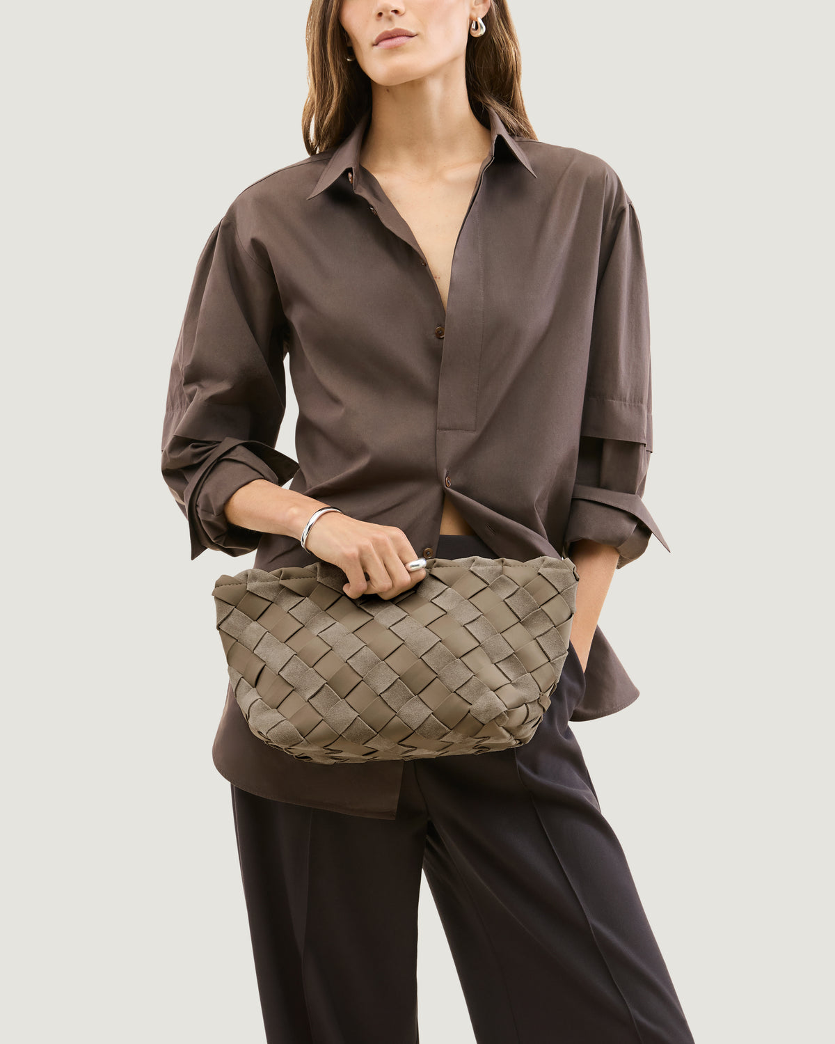 Model holding a handwoven suede and neoprene clutch bag in a taupe-beige color, featuring a basketweave pattern and a top zipper closure.