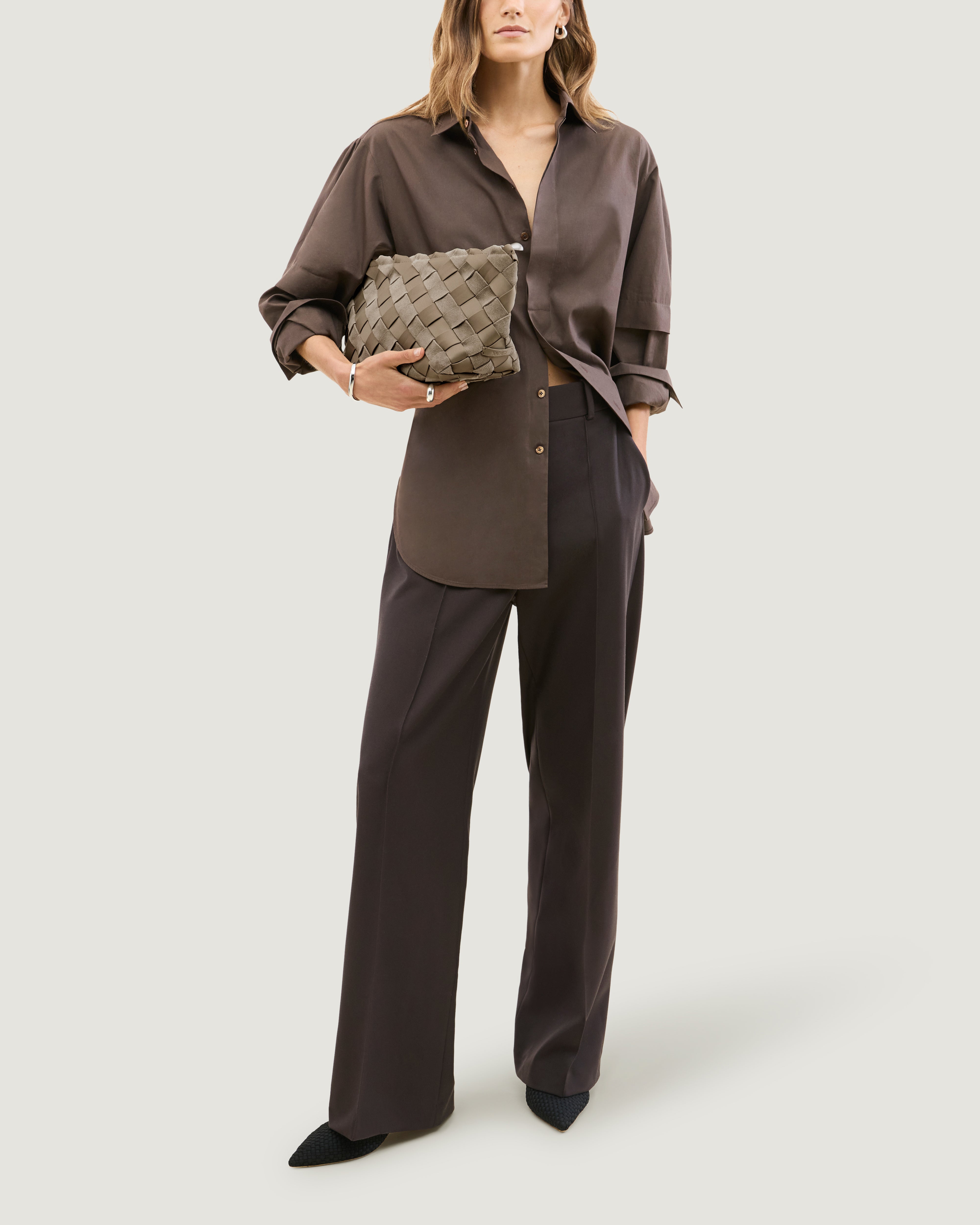 Model holding a handwoven suede and neoprene clutch bag in a taupe-beige color under her arm.