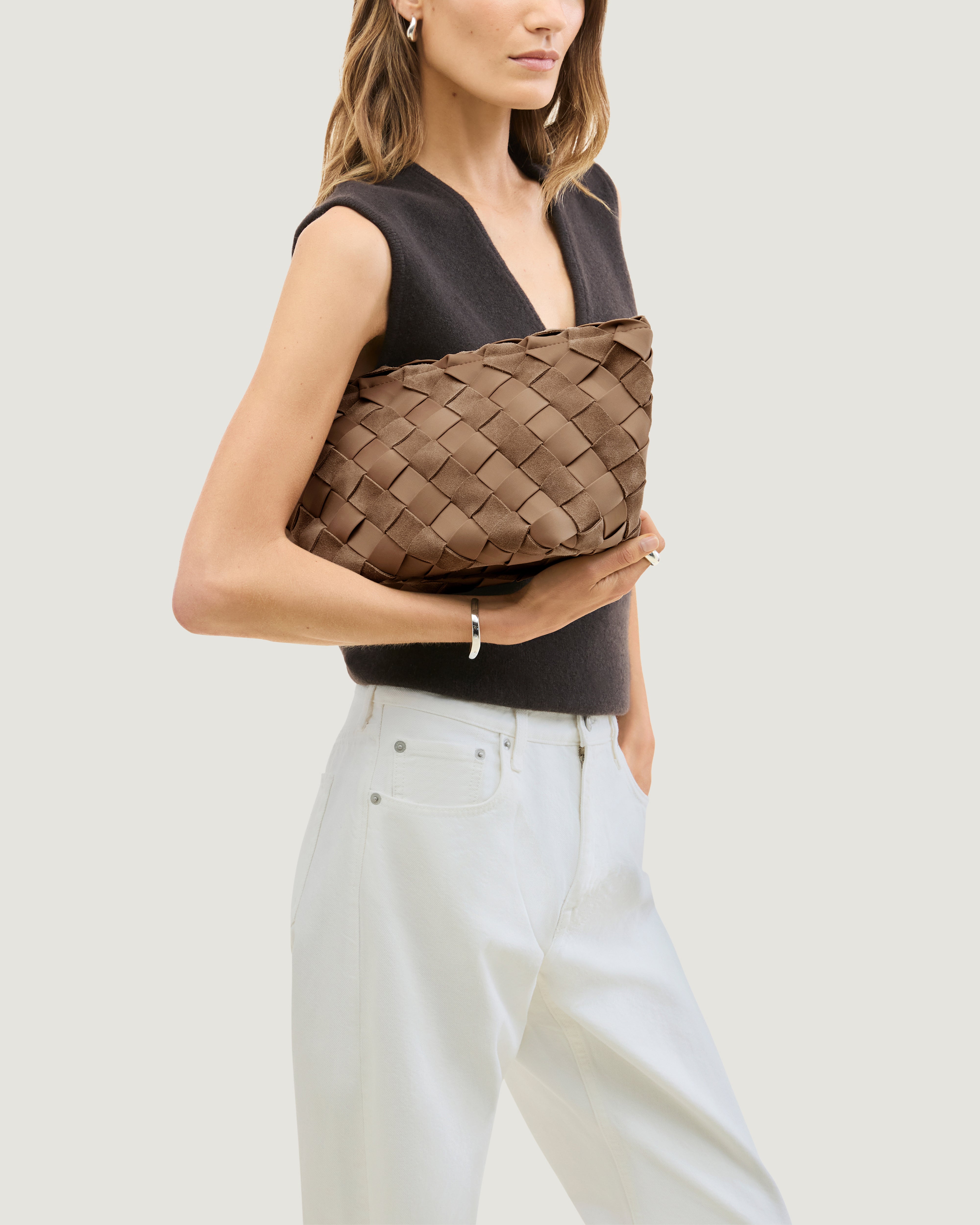 A model carrying a large handwoven suede and neoprene pouch in a medium brown shade, featuring a top zipper closure.