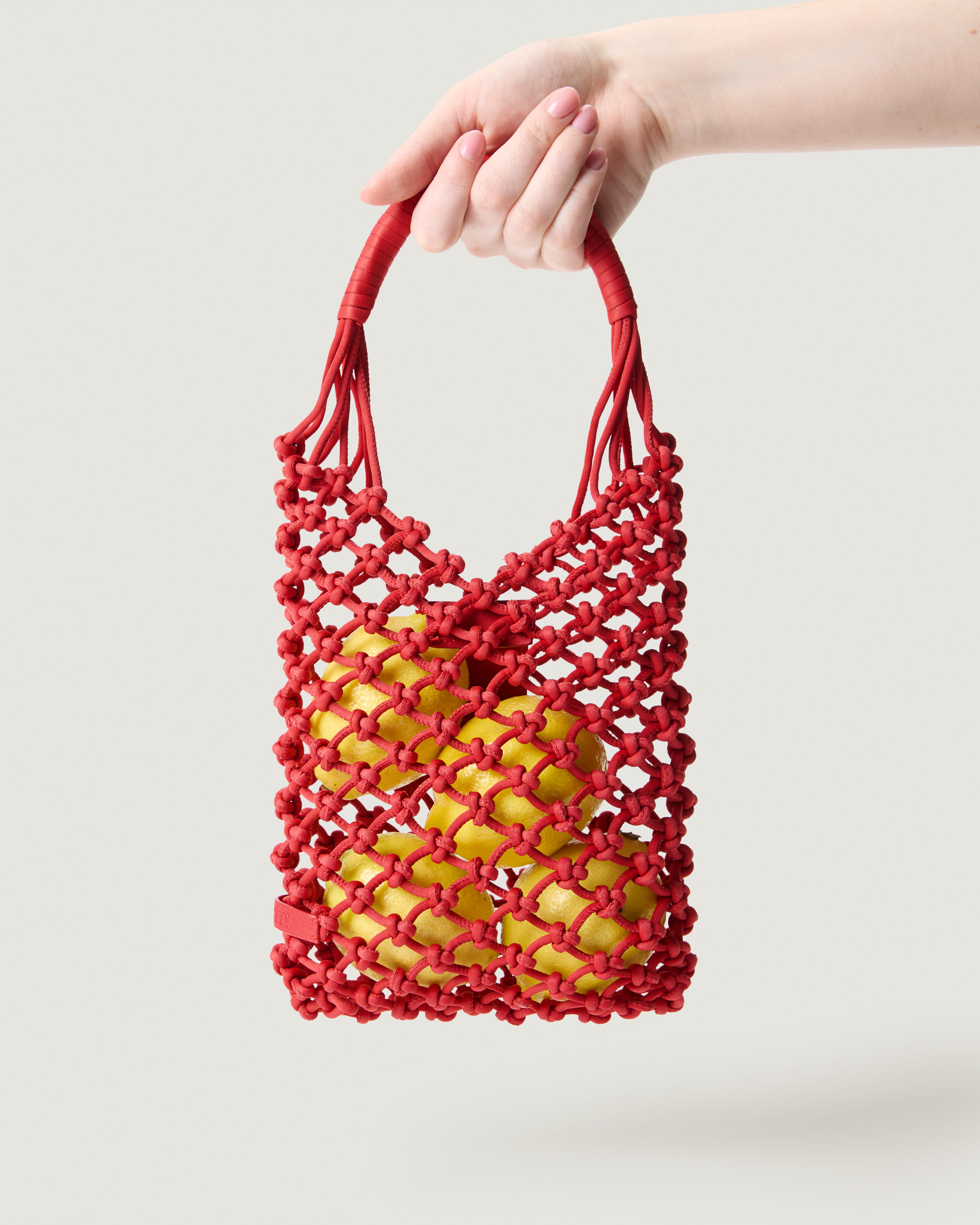 A hand holding small woven neoprene net bag in cherry red with fruit inside