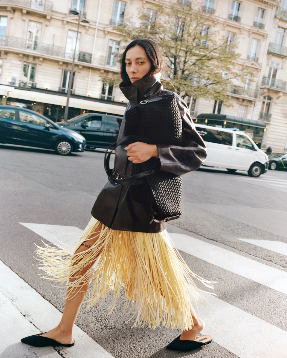 Close-up of a large black woven neoprene weekender bag with leather shoulder straps, carried by a model on a city street.