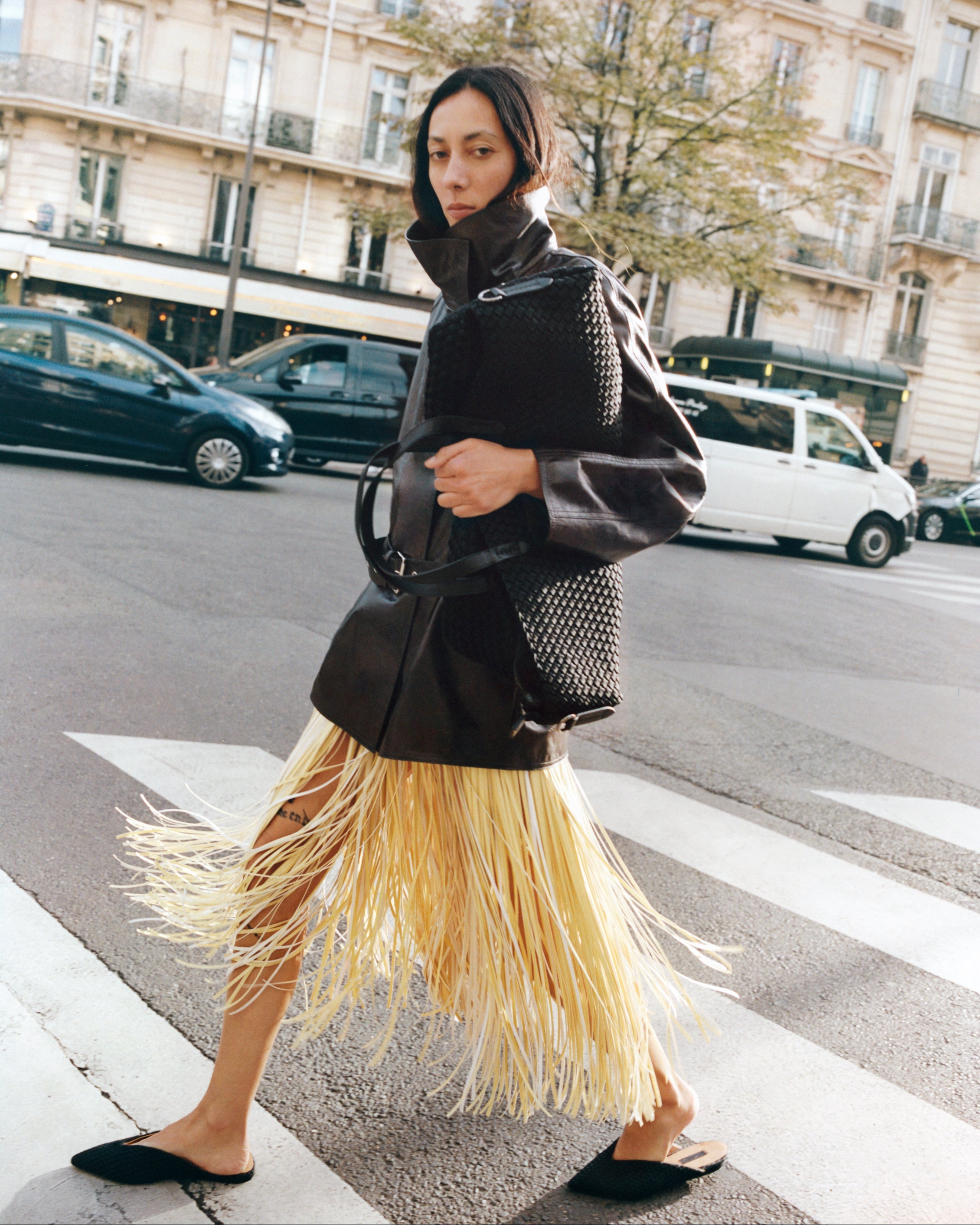 Close-up of a large black woven neoprene weekender bag with leather shoulder straps, carried by a model on a city street.