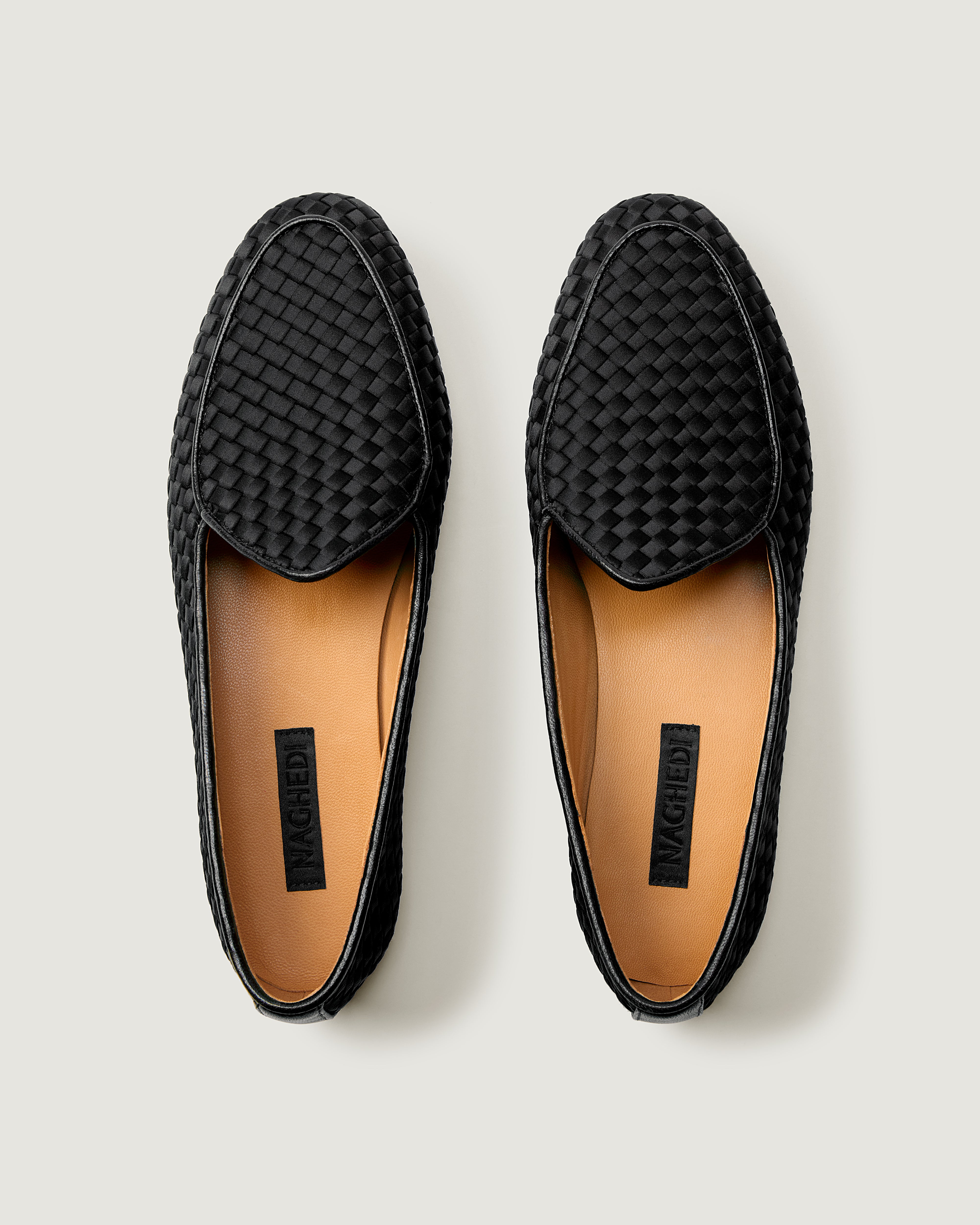 A pair of black handwoven satin slip-on loafers shown from a top-down view on a light gray background. The shoes feature a textured basketweave pattern on the vamp and a tan interior.