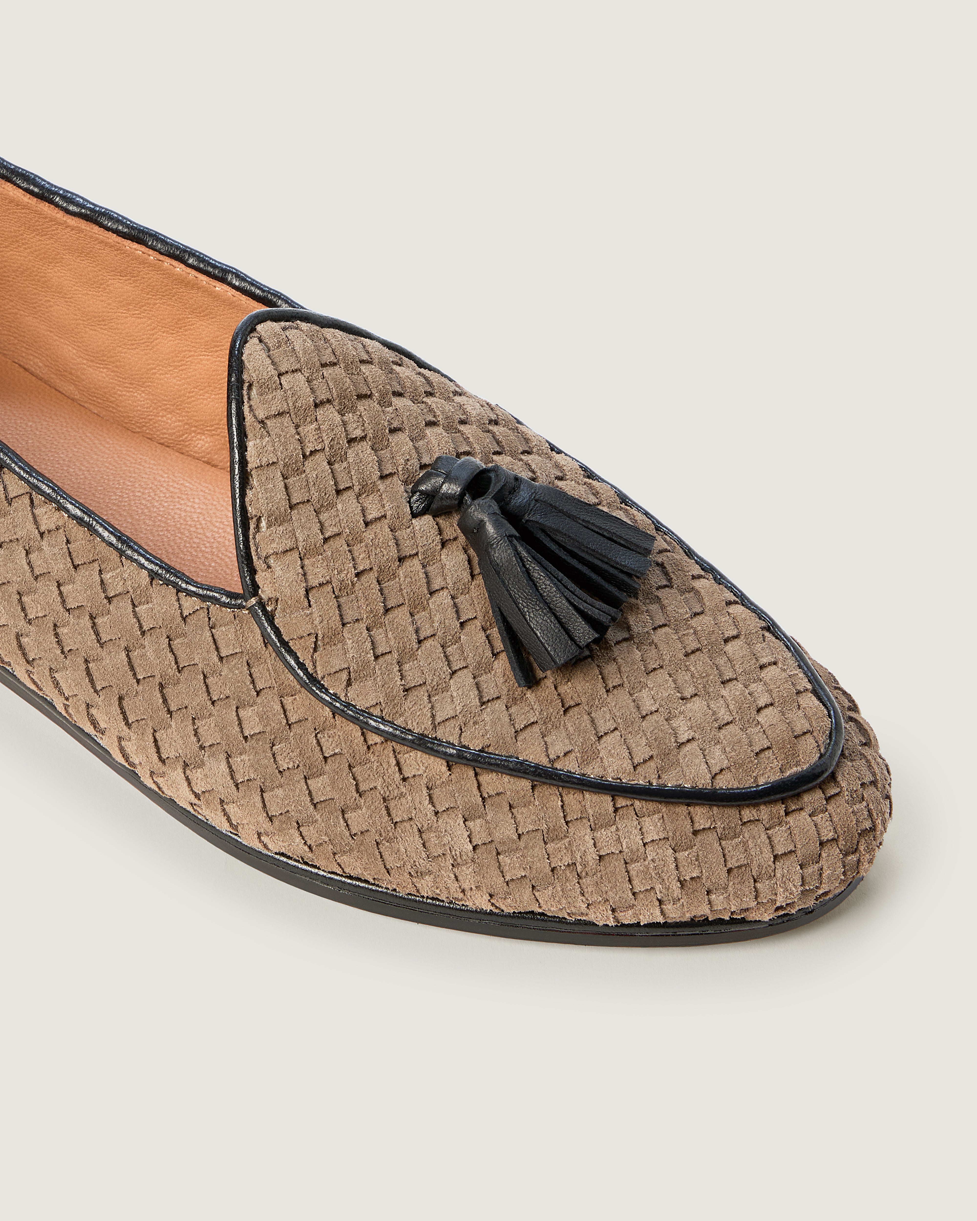 A close up of single handwoven suede loafer in beige with a black tassel and black trim against a light gray background.