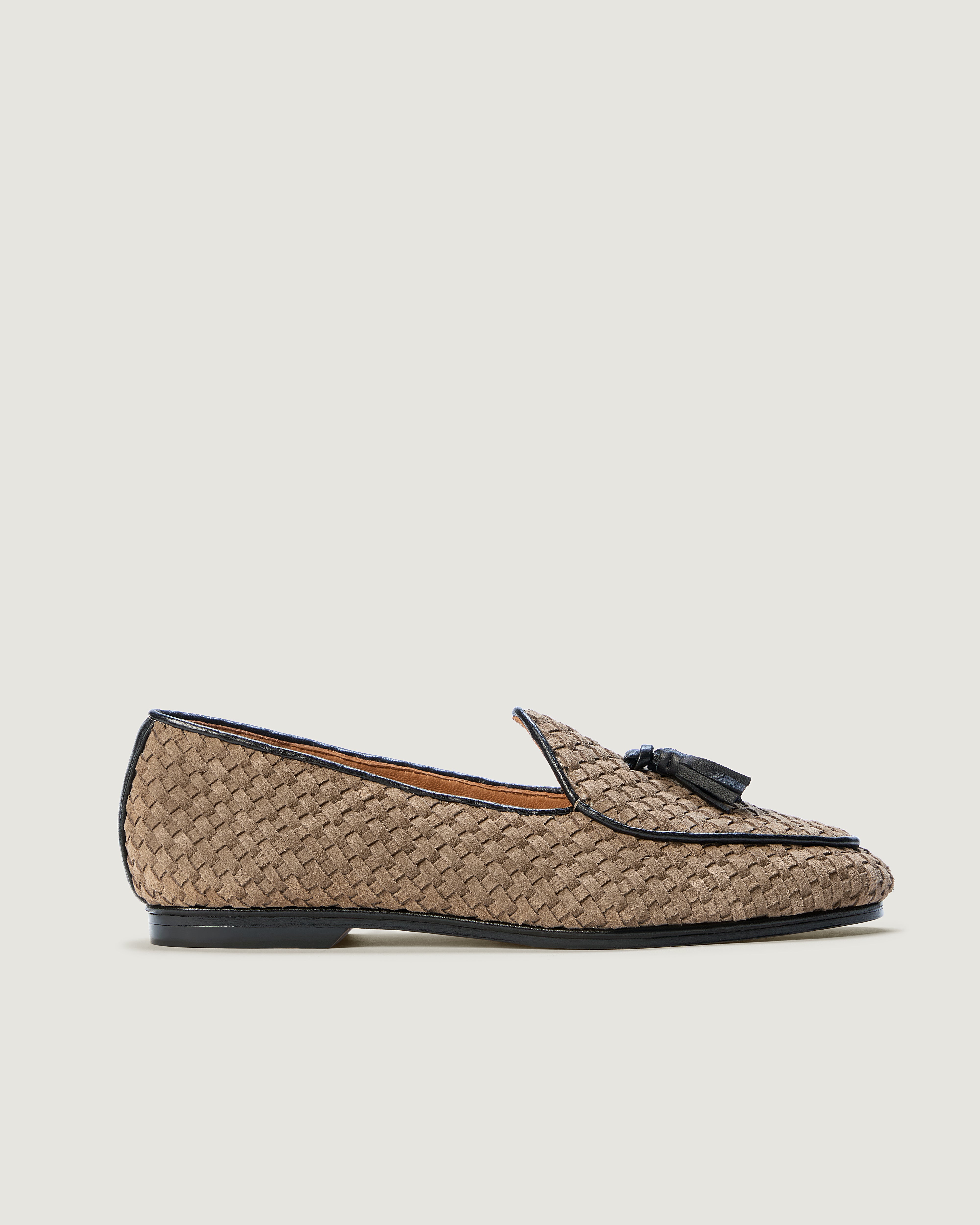 A single handwoven suede loafer in beige with a black tassel and black trim, shown in side profile against a light gray background.