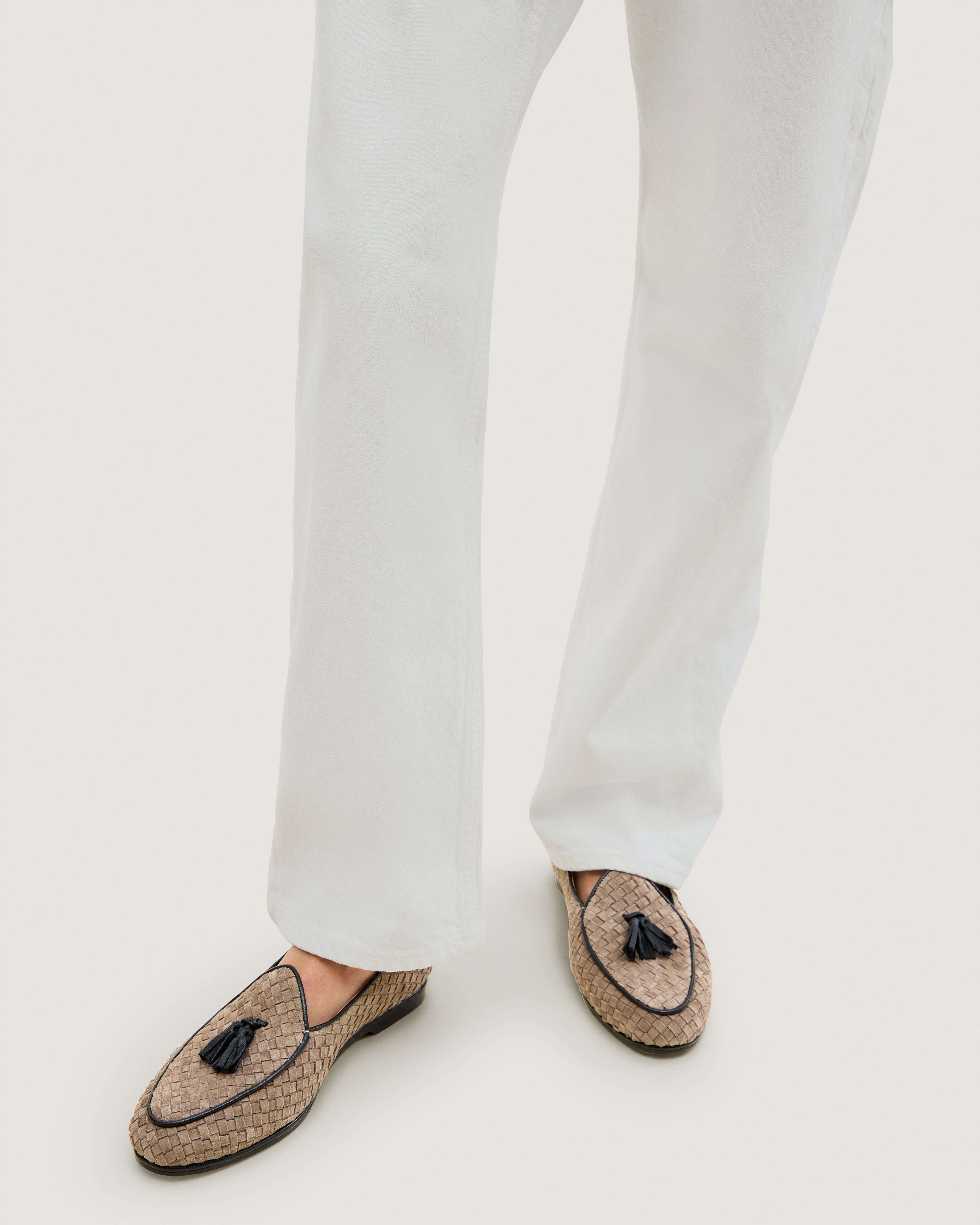 Model wearing white pants and a pair of suede loafers in beige with a black tassel and black trim.