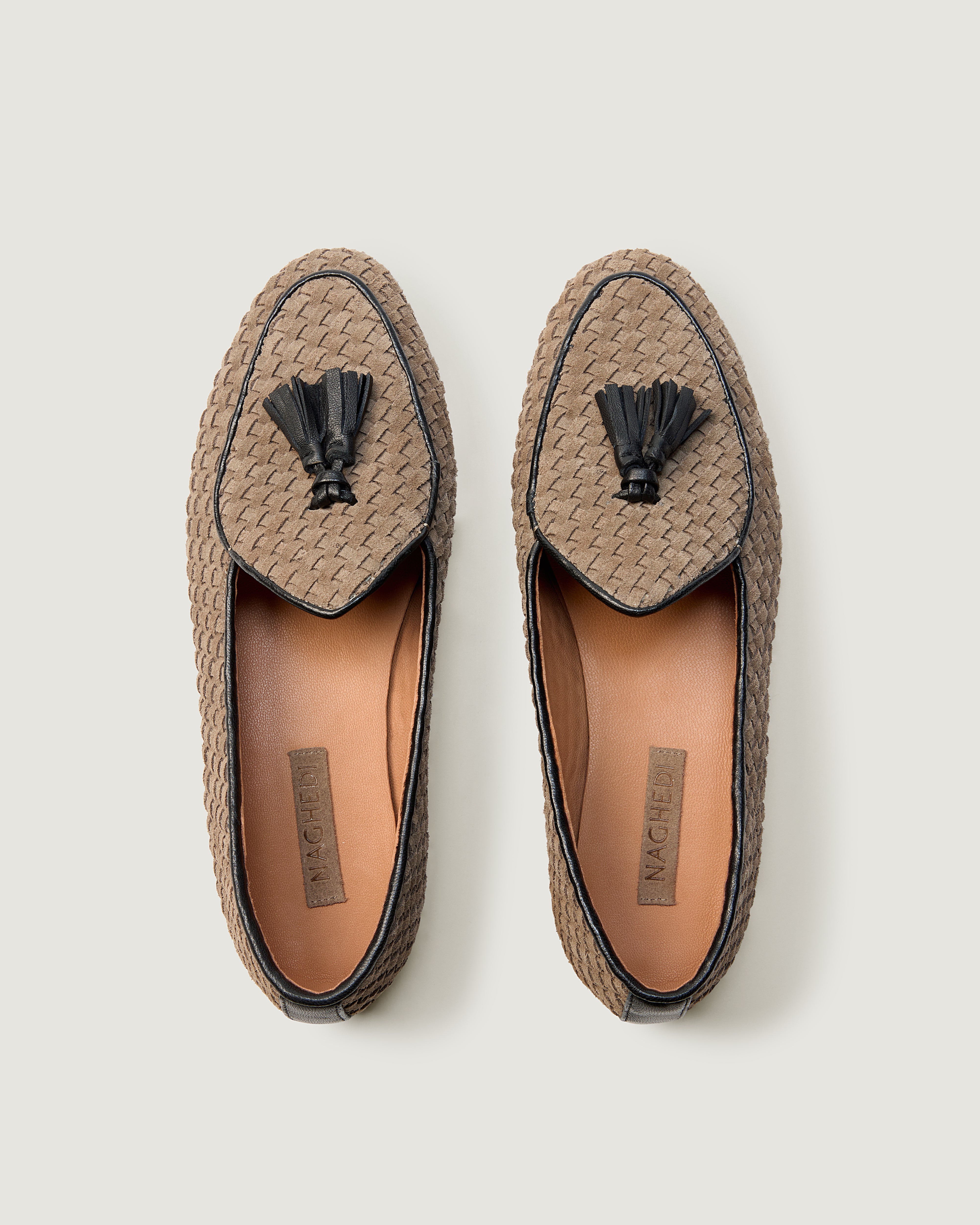A top view of a pair of suede loafers in beige with a black tassel and black trim, shown in side profile against a light gray background.