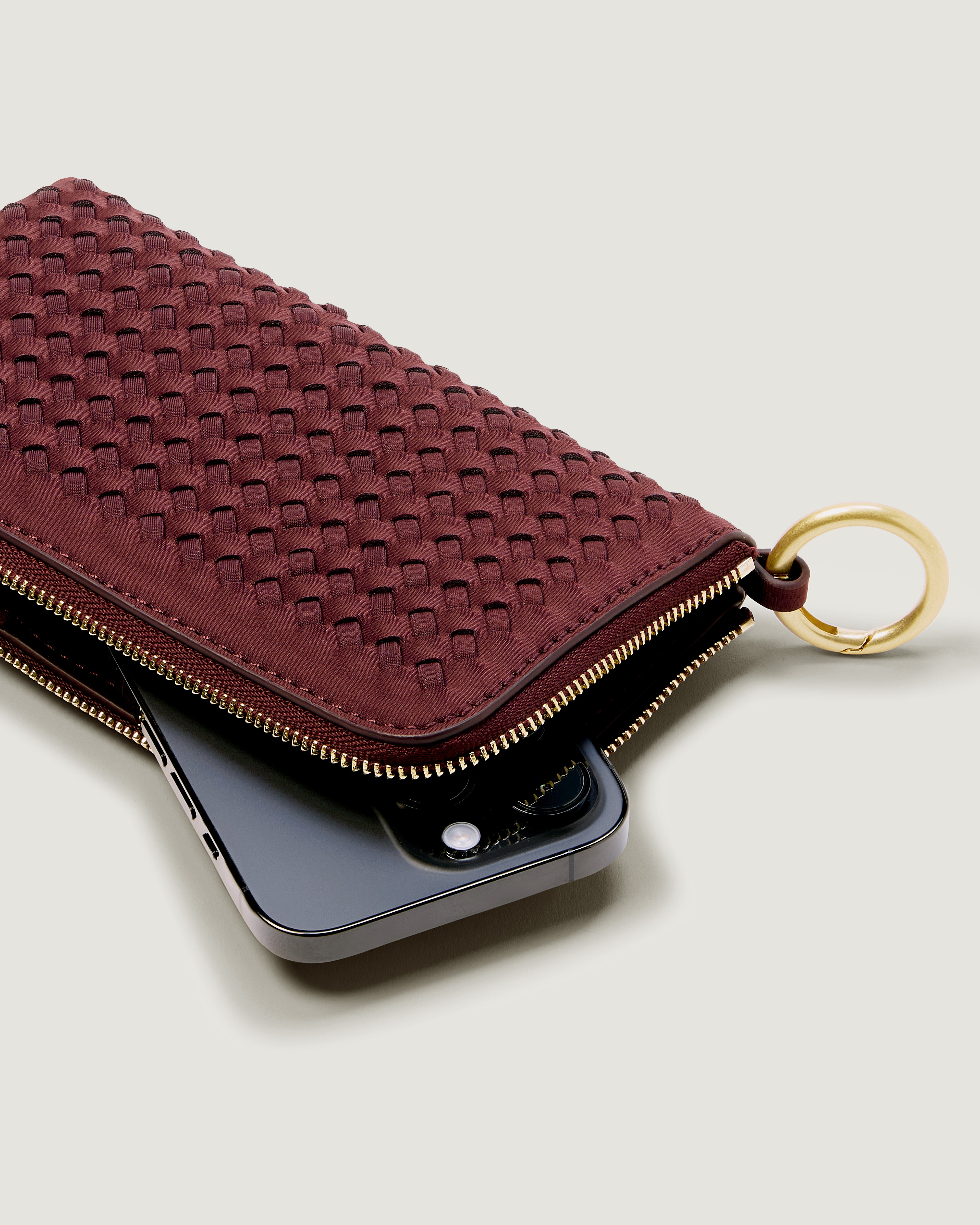 A handwoven neoprene crossbody zippered phone case in a burgundy color, with an iPhone inside.