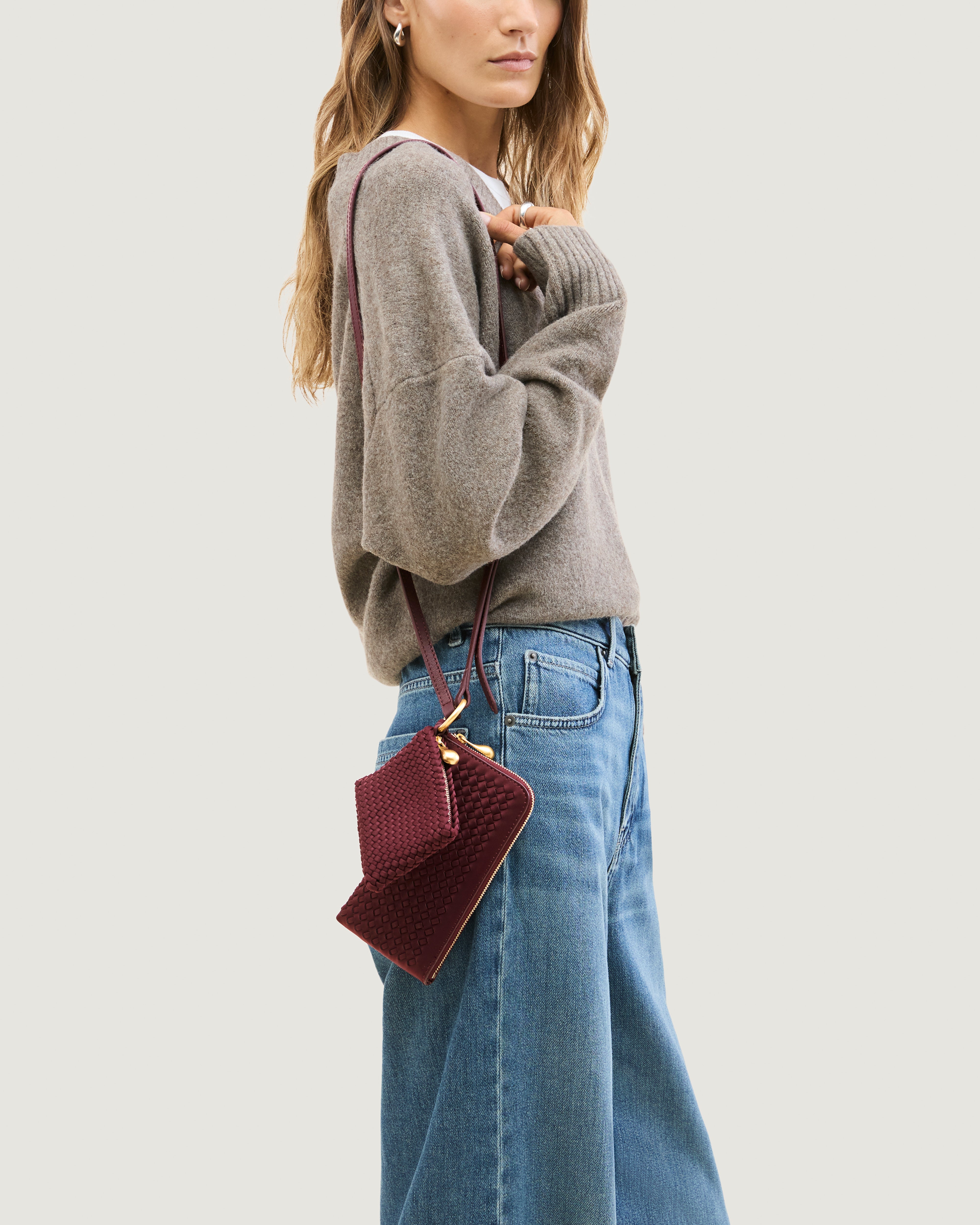 Model wearing a handwoven neoprene crossbody zippered phone case in a burgundy color, featuring a matching zippered card holder pouch and a thin adjustable shoulder strap.