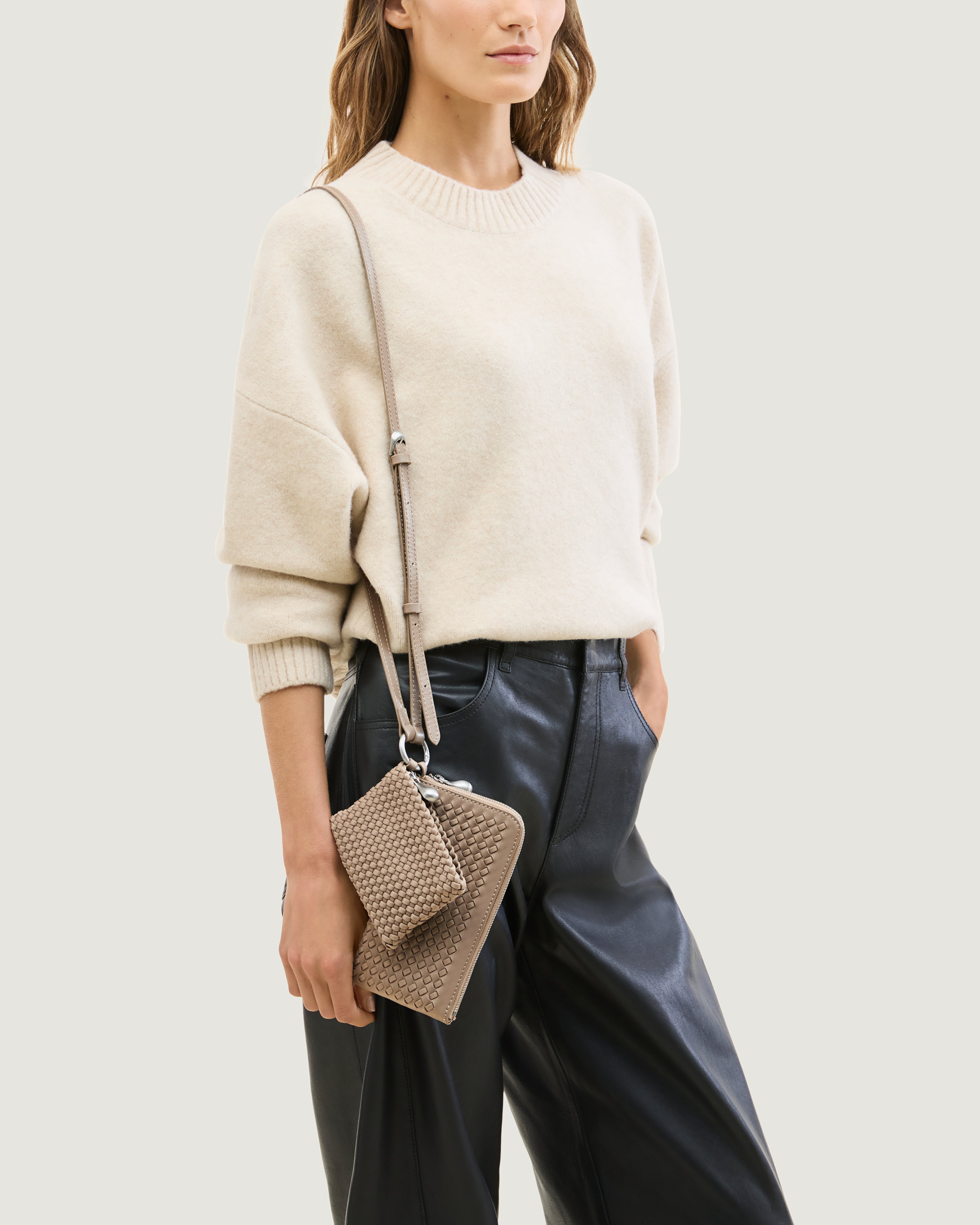 Model wearing a handwoven neoprene crossbody zippered phone case in a taupe-brown color, featuring a matching zippered card holder pouch and a thin adjustable shoulder strap.