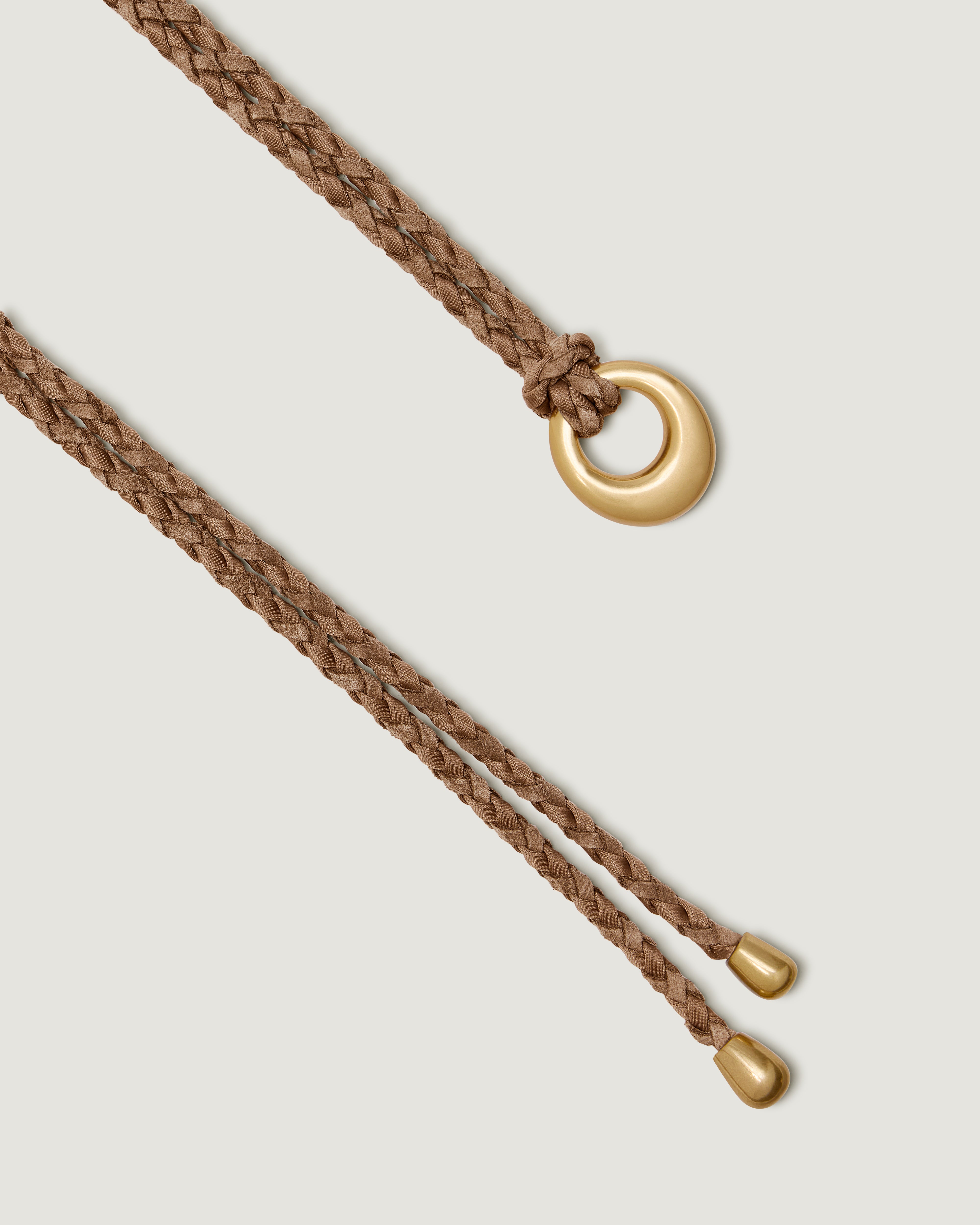 A close up of the gold metal ring closure and matching gold-toned tips on a light brown handwoven neoprene and suede braided rope belt.