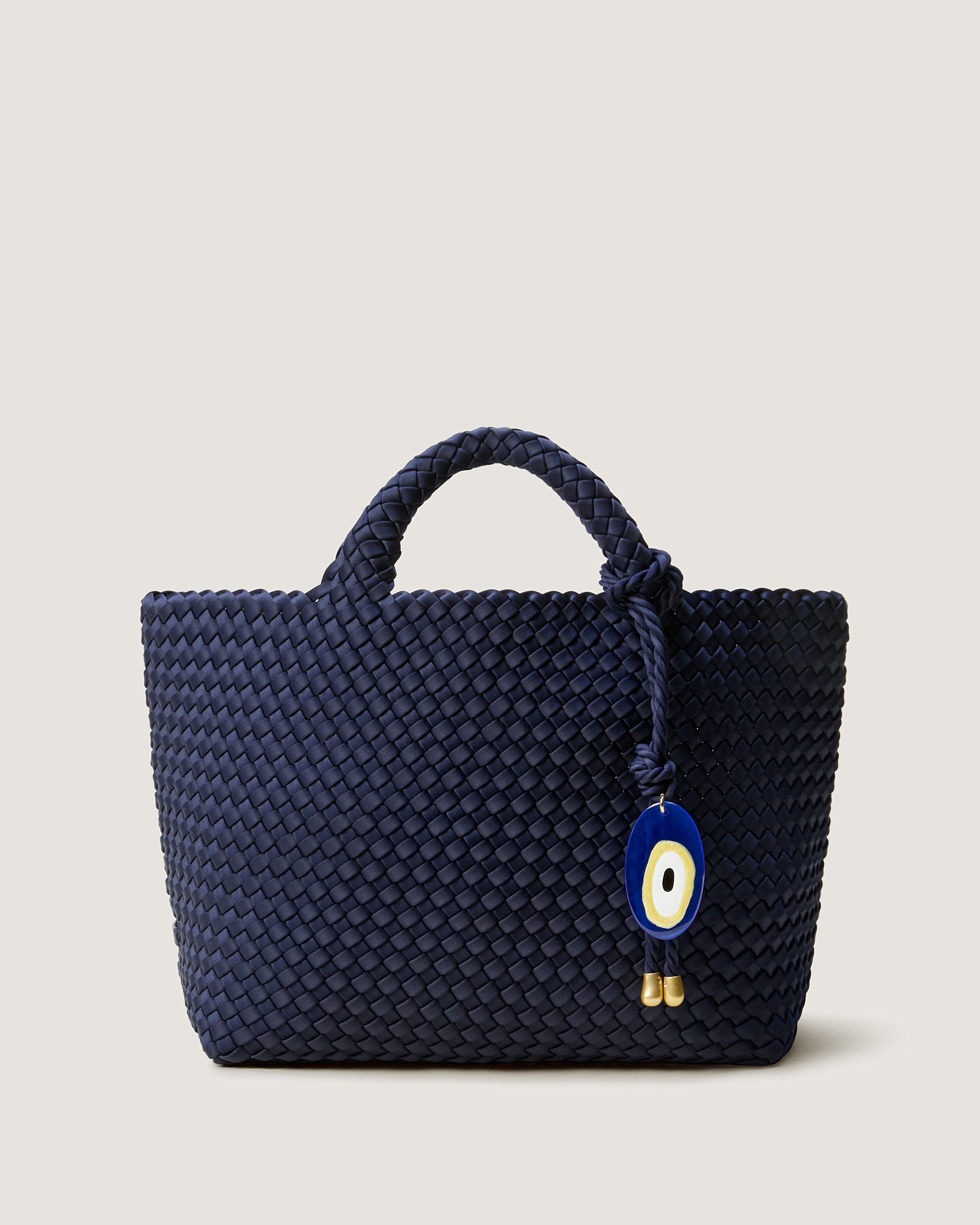 St. Barths Tote in Ink Blue, a dark navy blue tone, in handwoven neoprene, featuring an open top, and woven top handles with a matching Evil Eye rope bag charm.