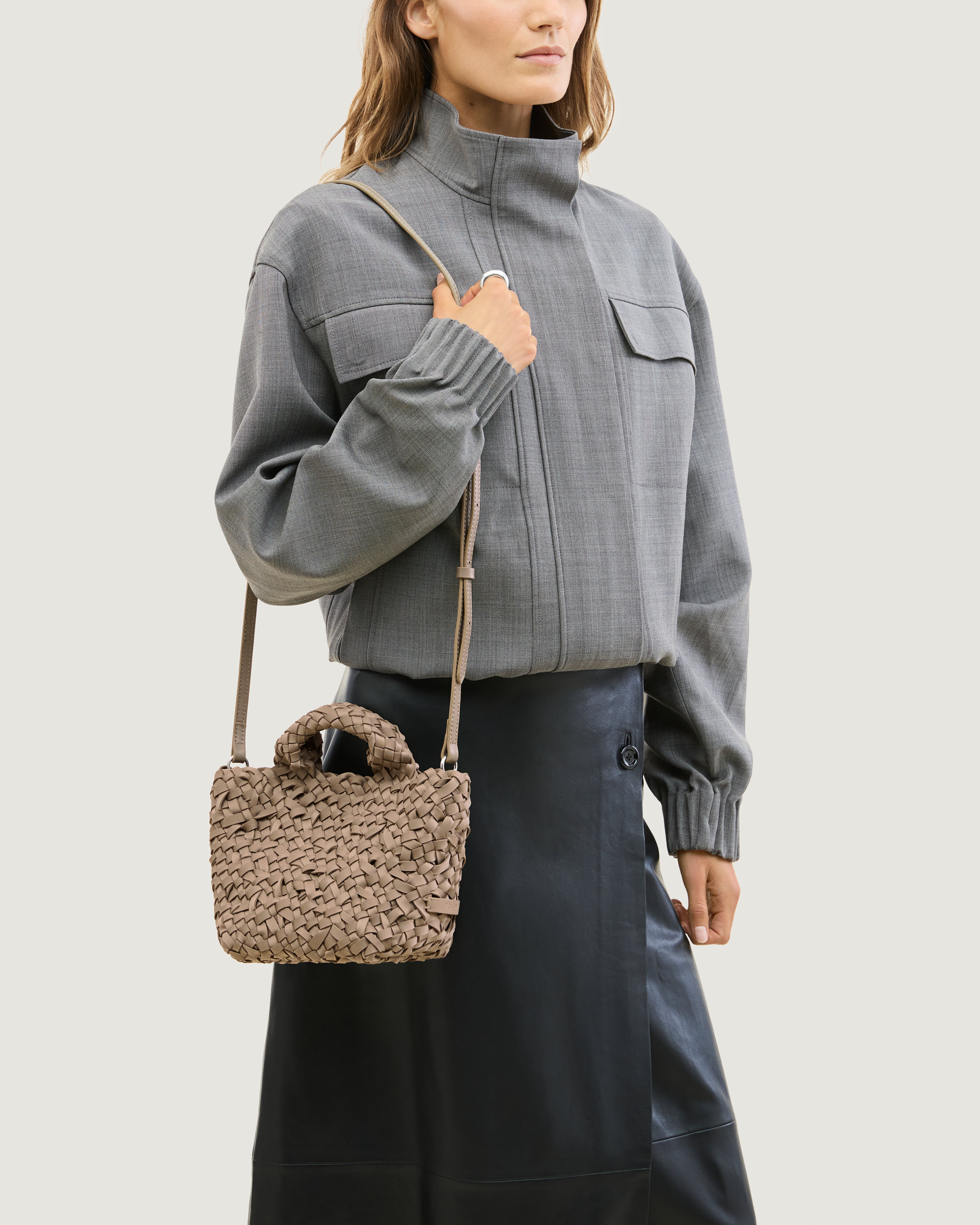 Model wearing a cool taupe woven basket-style neoprene handbag with a top handle and a leather shoulder strap.