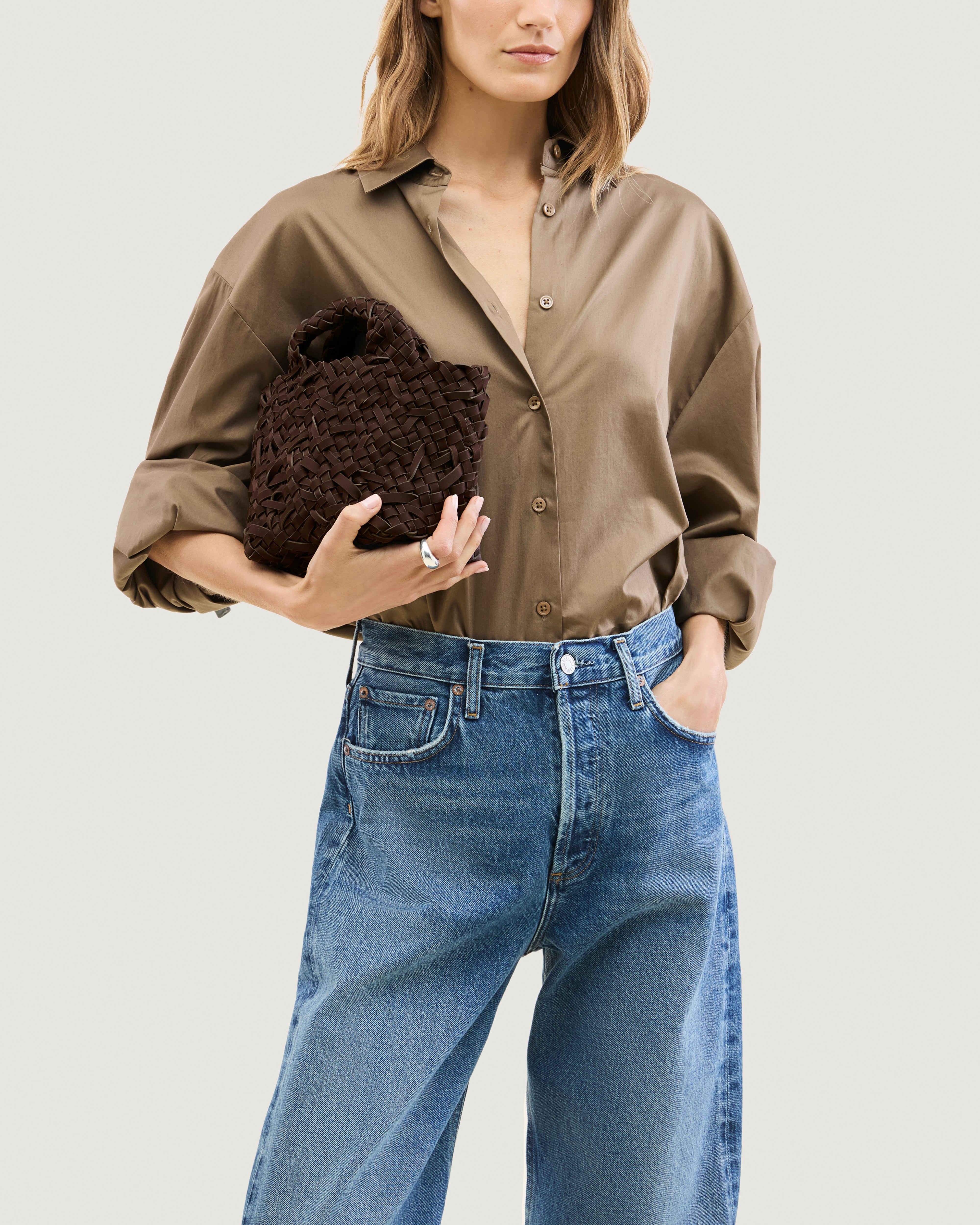 Model holding a chocolate brown woven basket-style neoprene handbag with a top handle.