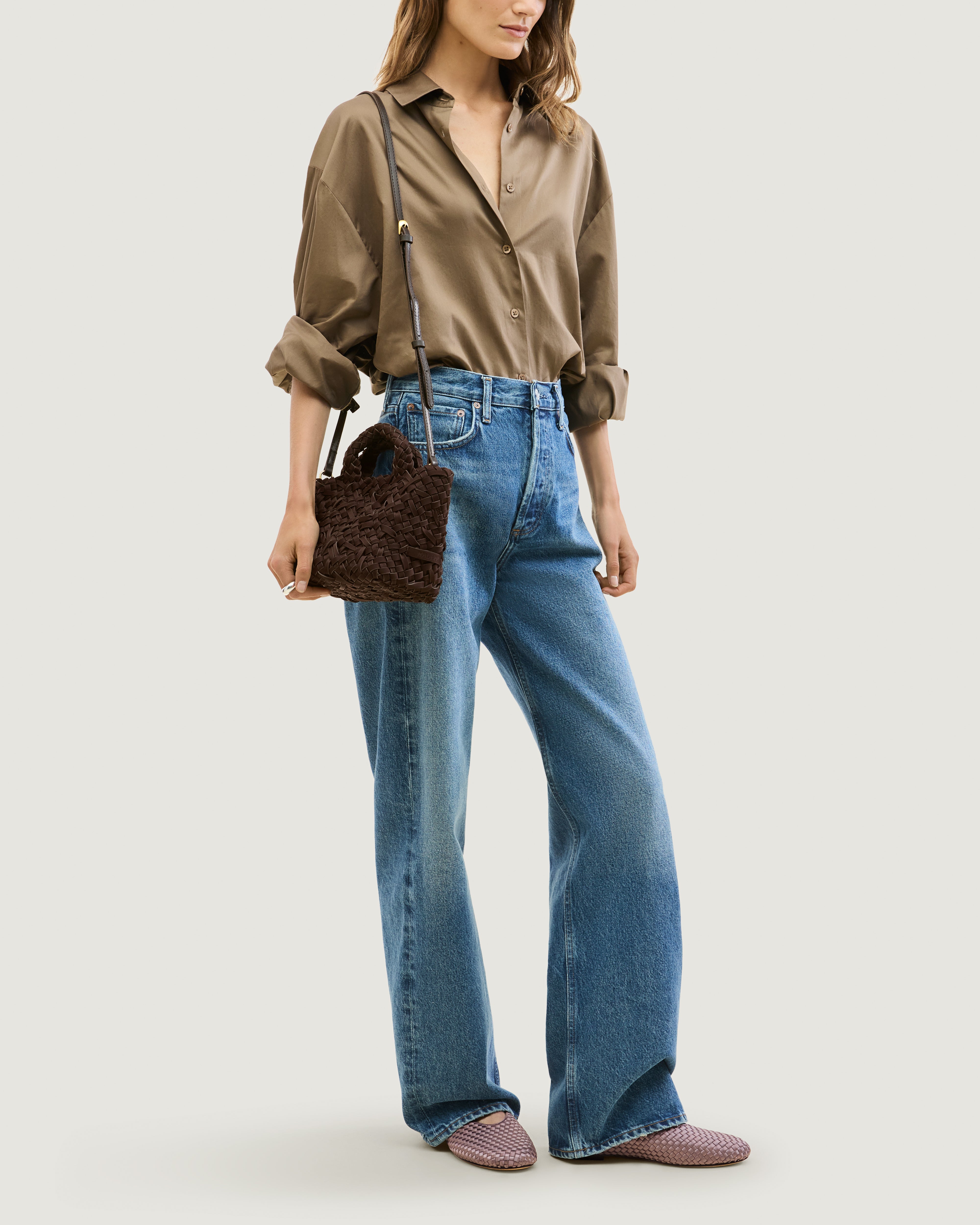 Model wearing a chocolate brown woven basket-style neoprene handbag with a top handle and a leather shoulder strap.